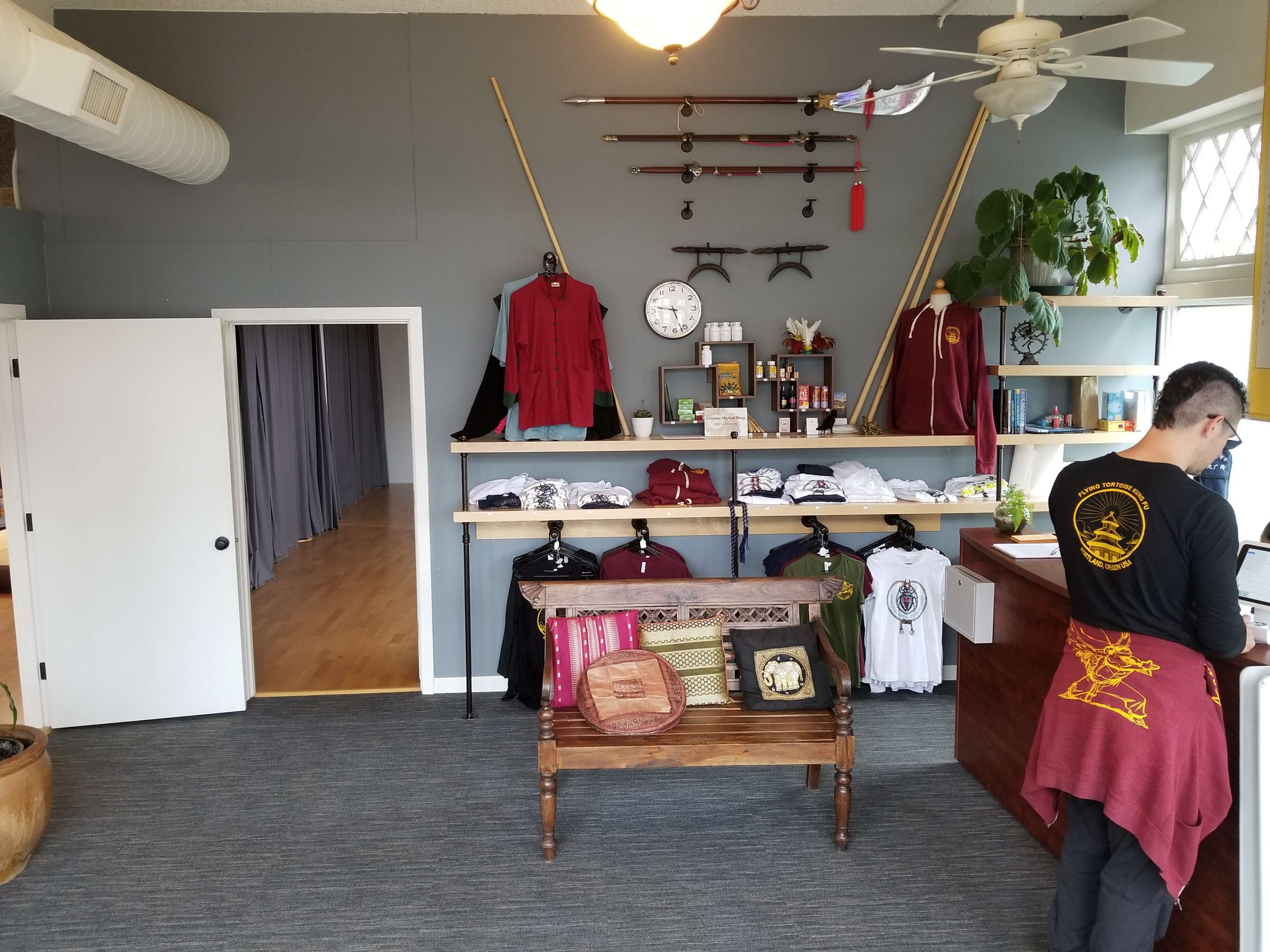 The interior of a retail shop featuring clothing displays on a wooden rack, wall-mounted swords, and a counter where a person is working.