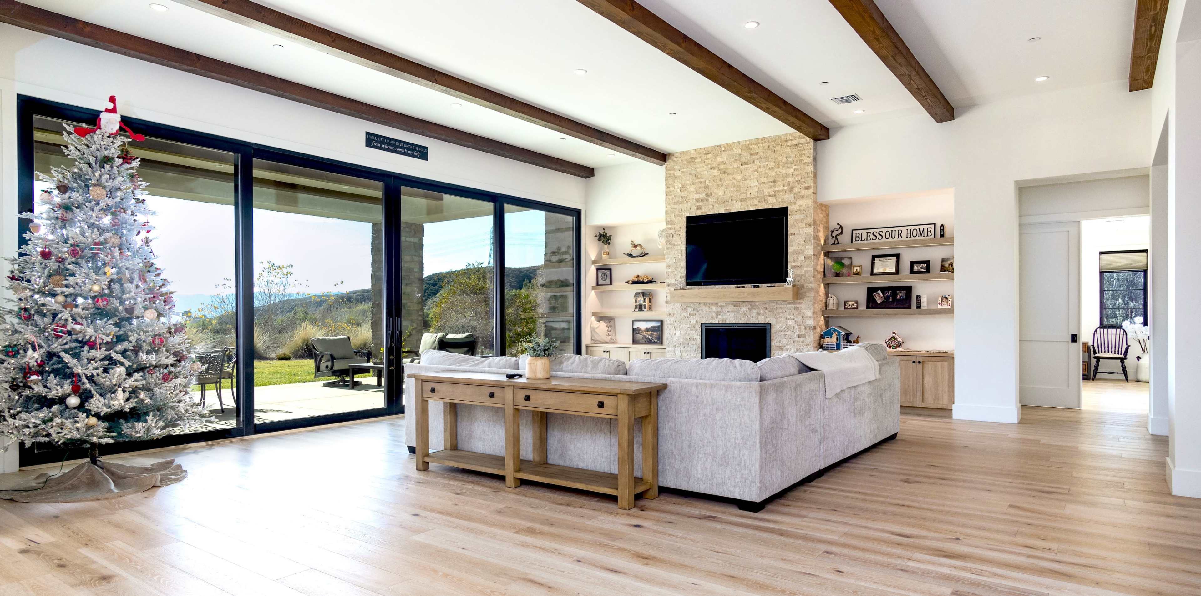 A modern living room features a large sliding glass door, a stone fireplace, a beige sofa, and a Christmas tree in the corner.