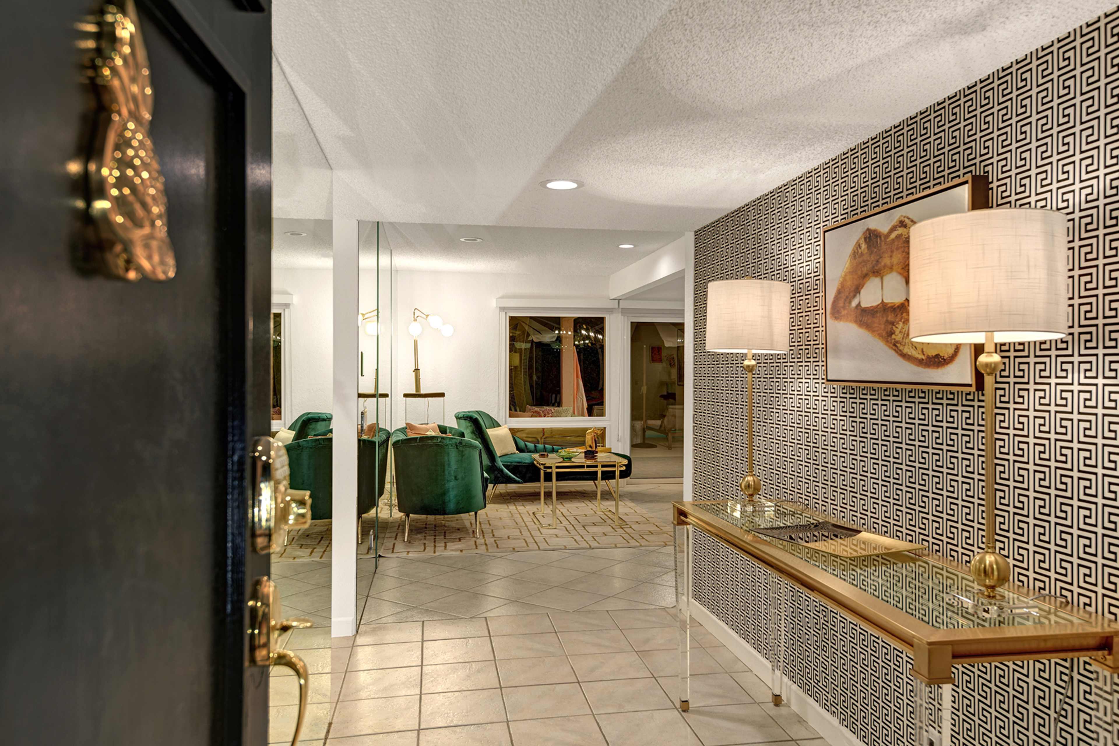 An entryway featuring a door with a decorative handle, a console table with a glass top, two lamps, and a patterned wall leading to a living area with green chairs.
