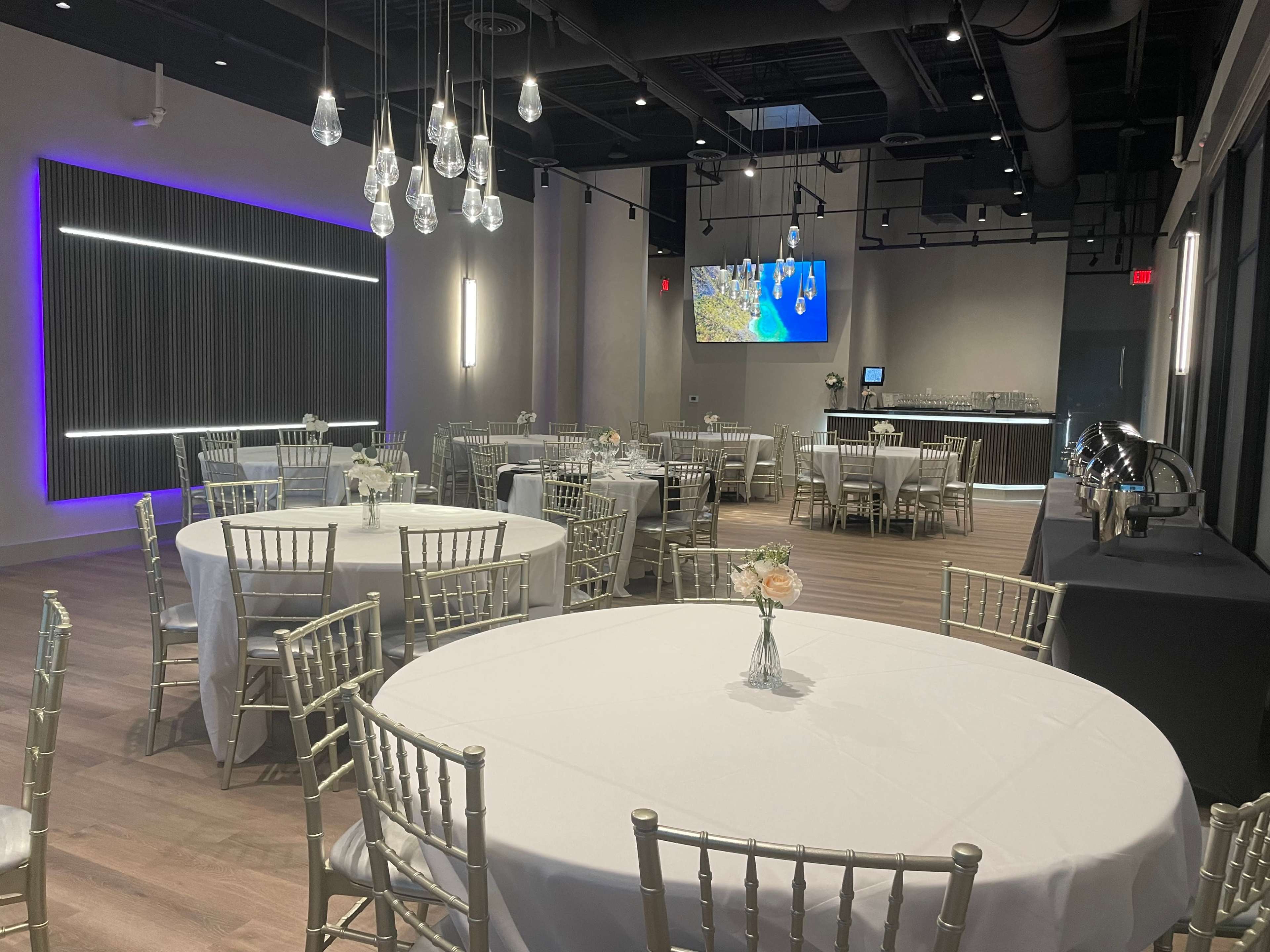 The interior of a modern event space features round tables with white tablecloths, silver chiavari chairs, and a bar area in the background with a large screen displaying a map.