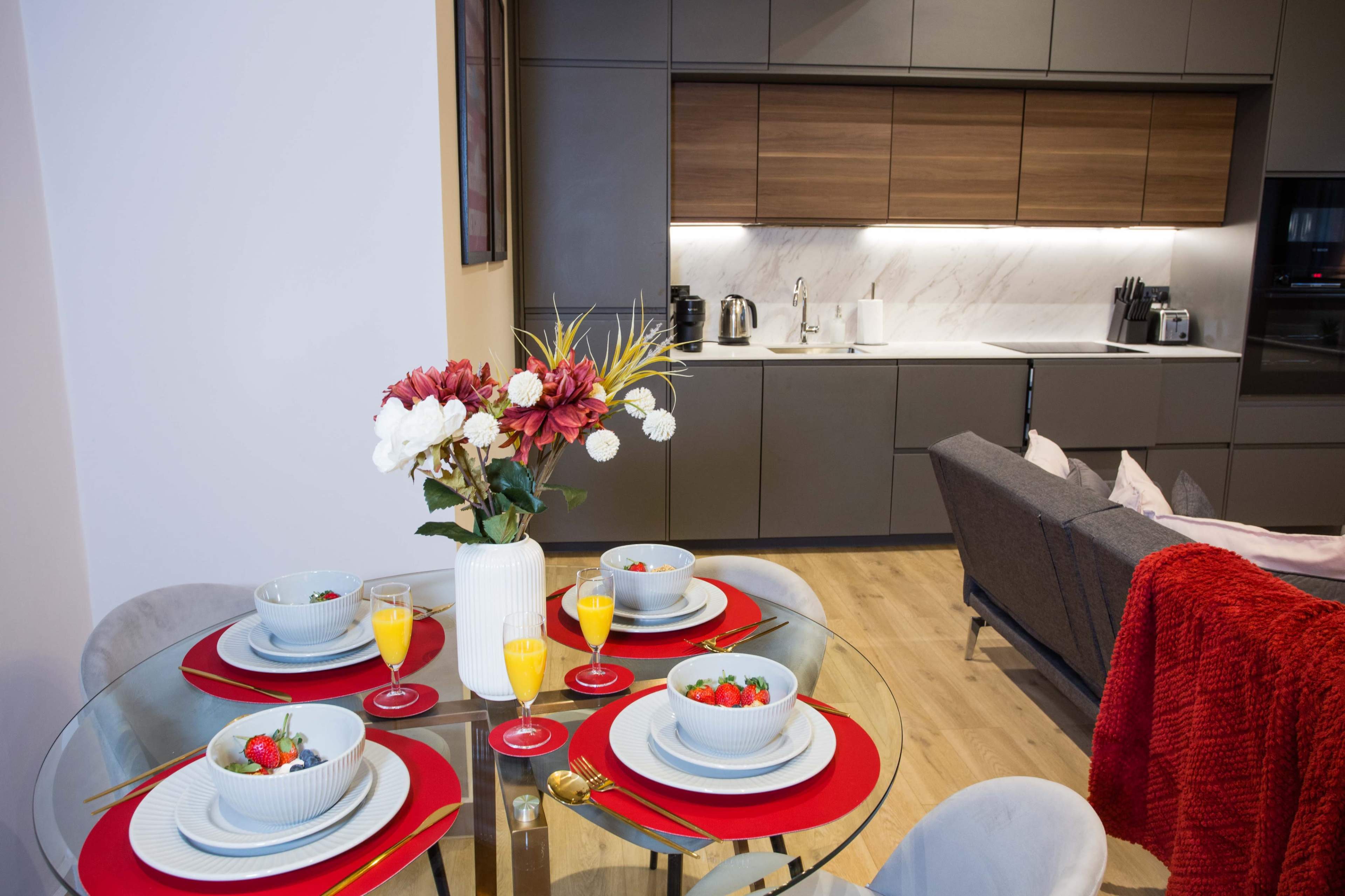 A dining table is set with bowls of fruit and drinks, while a modern kitchen with wood and marble finishes is visible in the background.