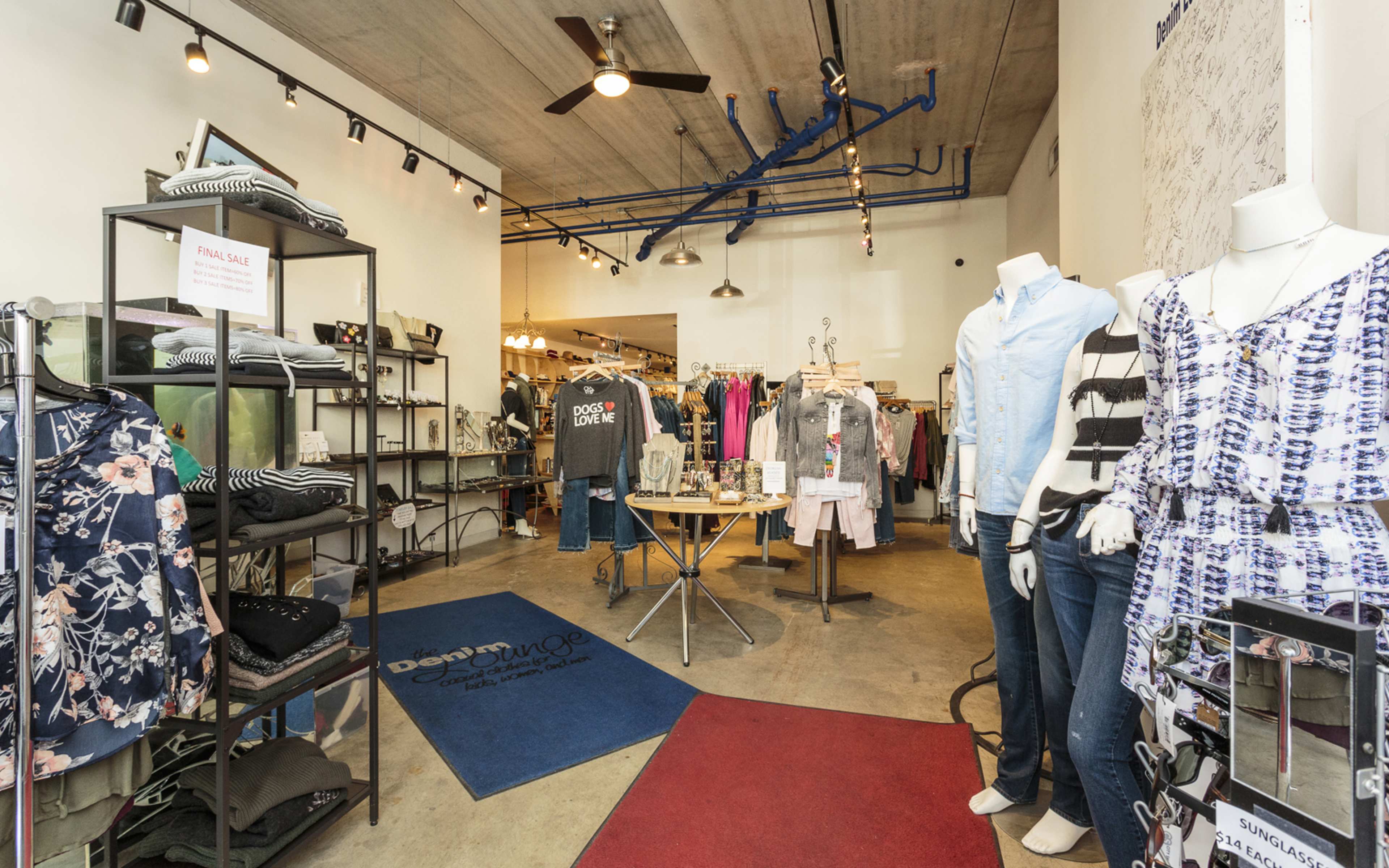 The image shows the interior of a clothing store with various garments displayed on racks and mannequins, featuring a blue and red entrance mat.