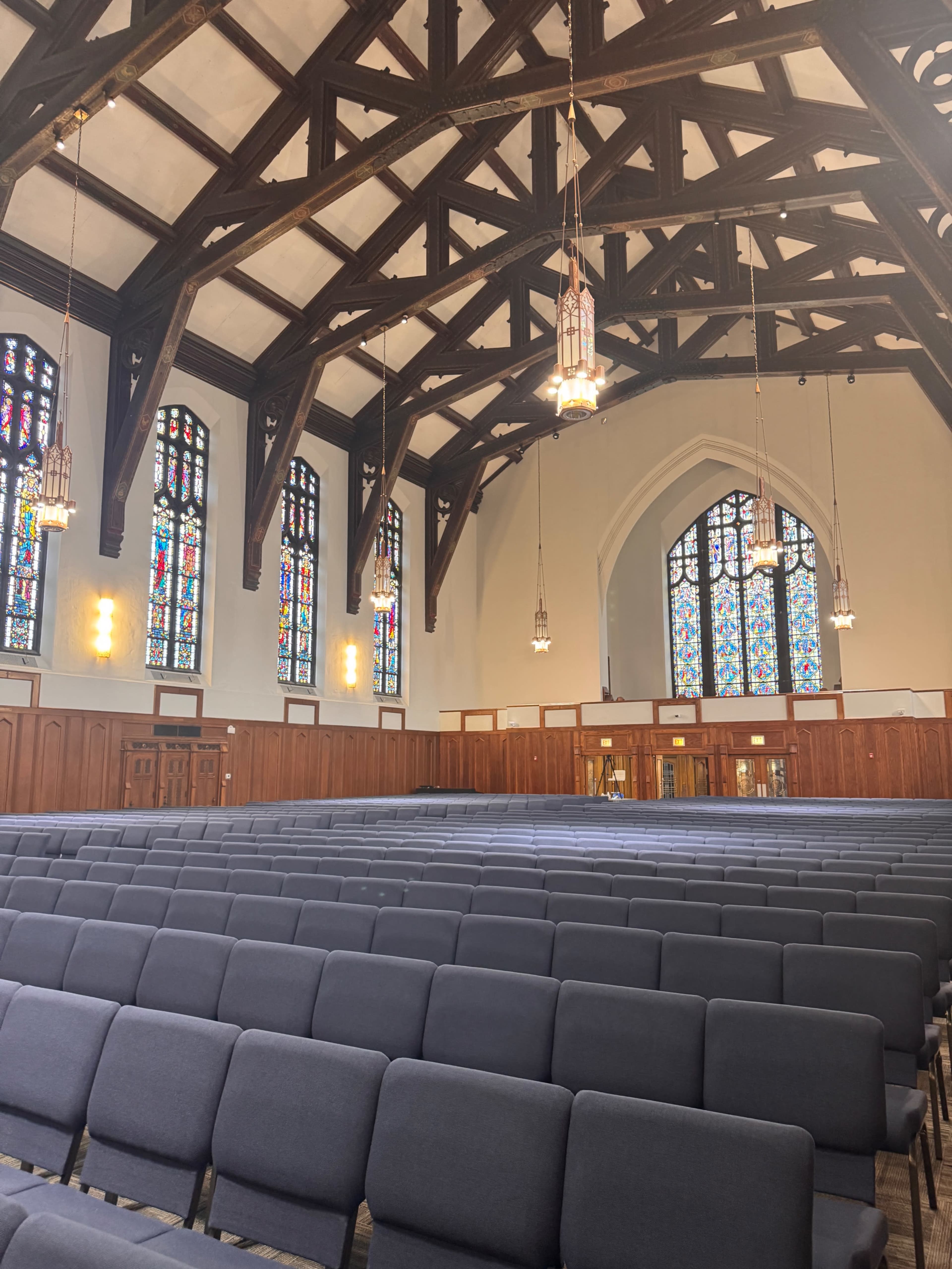 The image shows the interior of a church with wooden beams, stained glass windows, and rows of gray chairs facing the front.