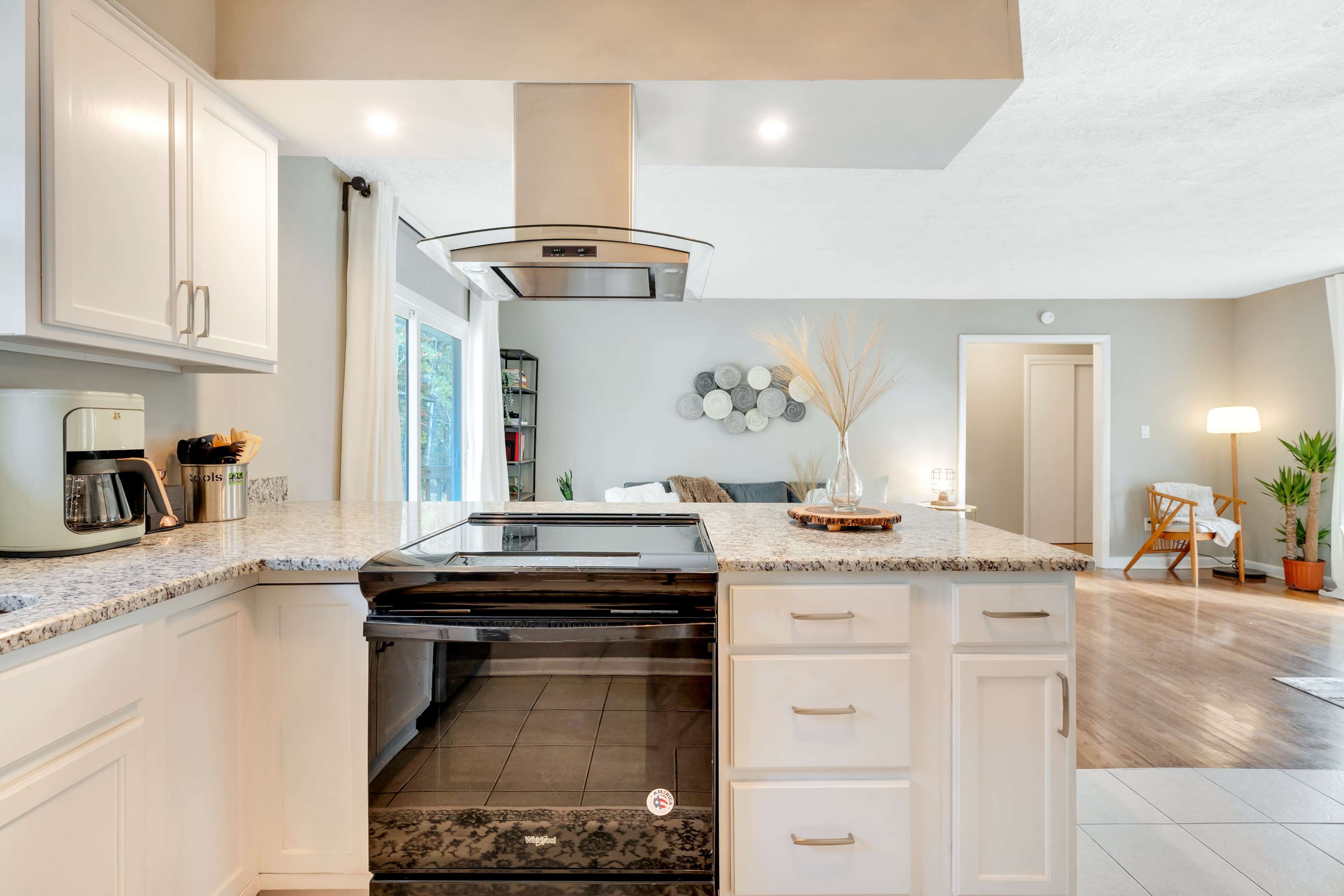 A modern kitchen features a central island with a stovetop, stainless steel hood, and an open layout connecting to a living area.