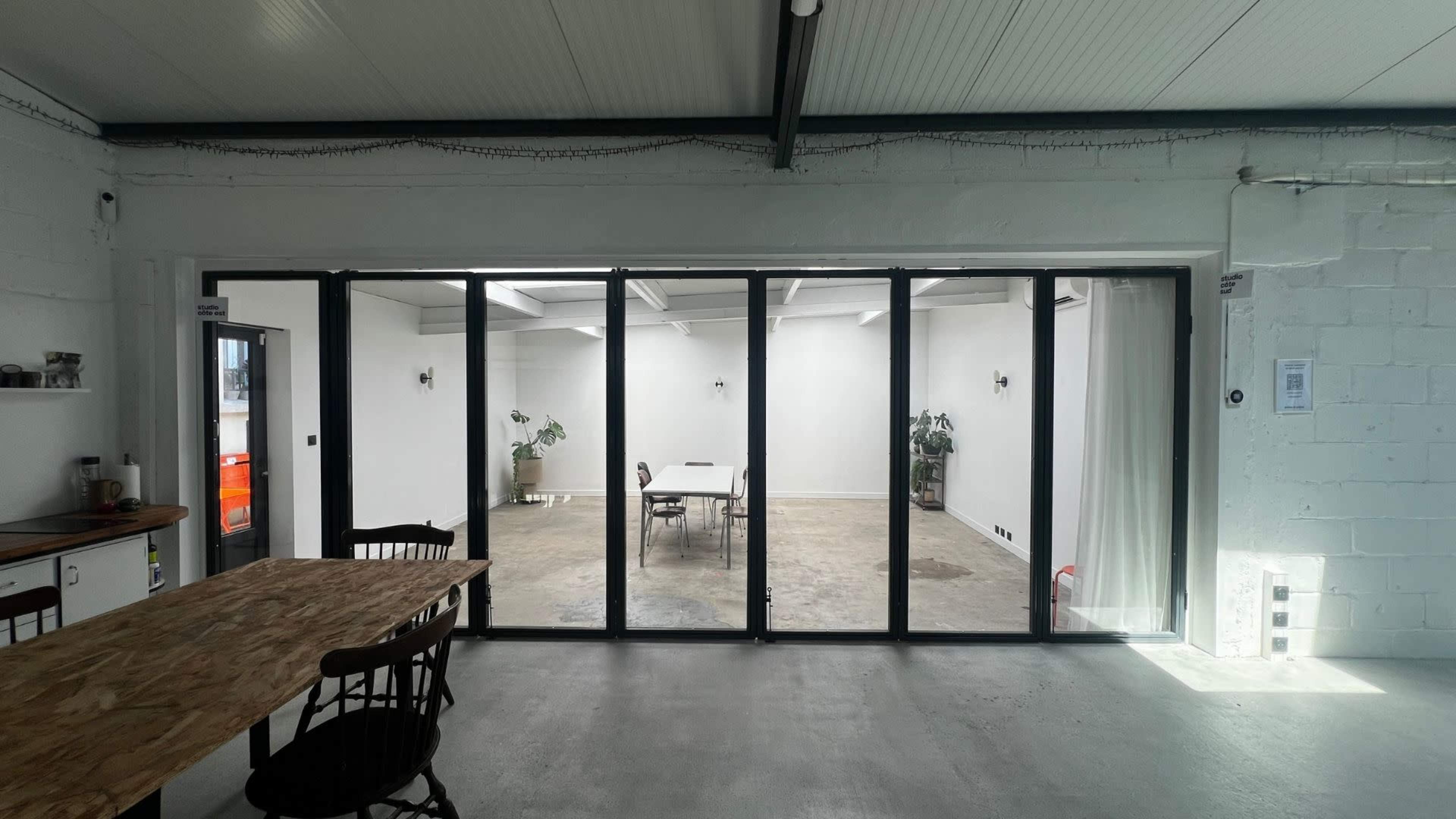 The image shows a bright, open room with large glass doors revealing a minimalist space featuring a table and chairs, along with potted plants.