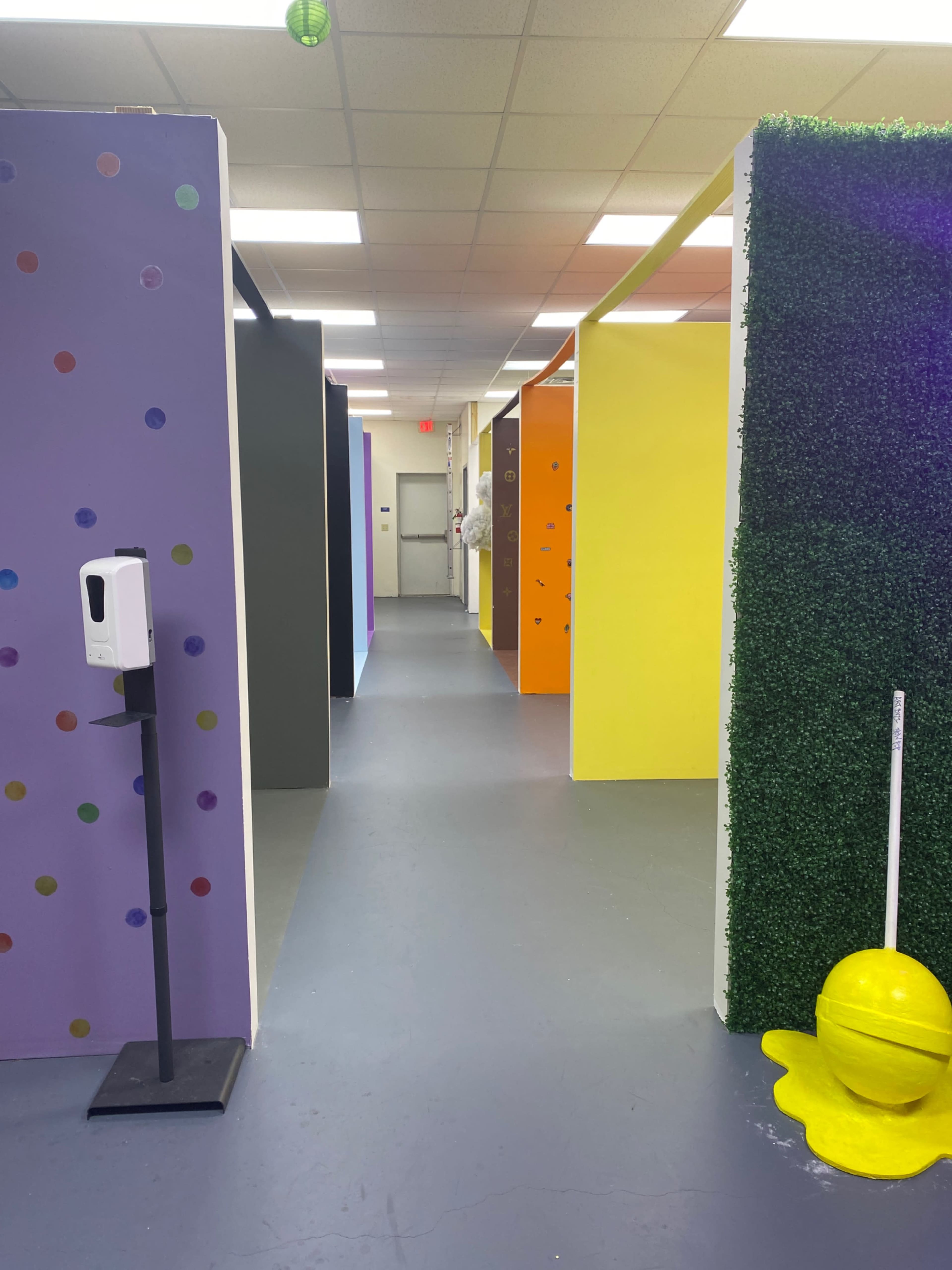 The image shows a hallway with colorful partitioned walls in various shades, including purple, yellow, and black, with a hand sanitizer station on the left side.
