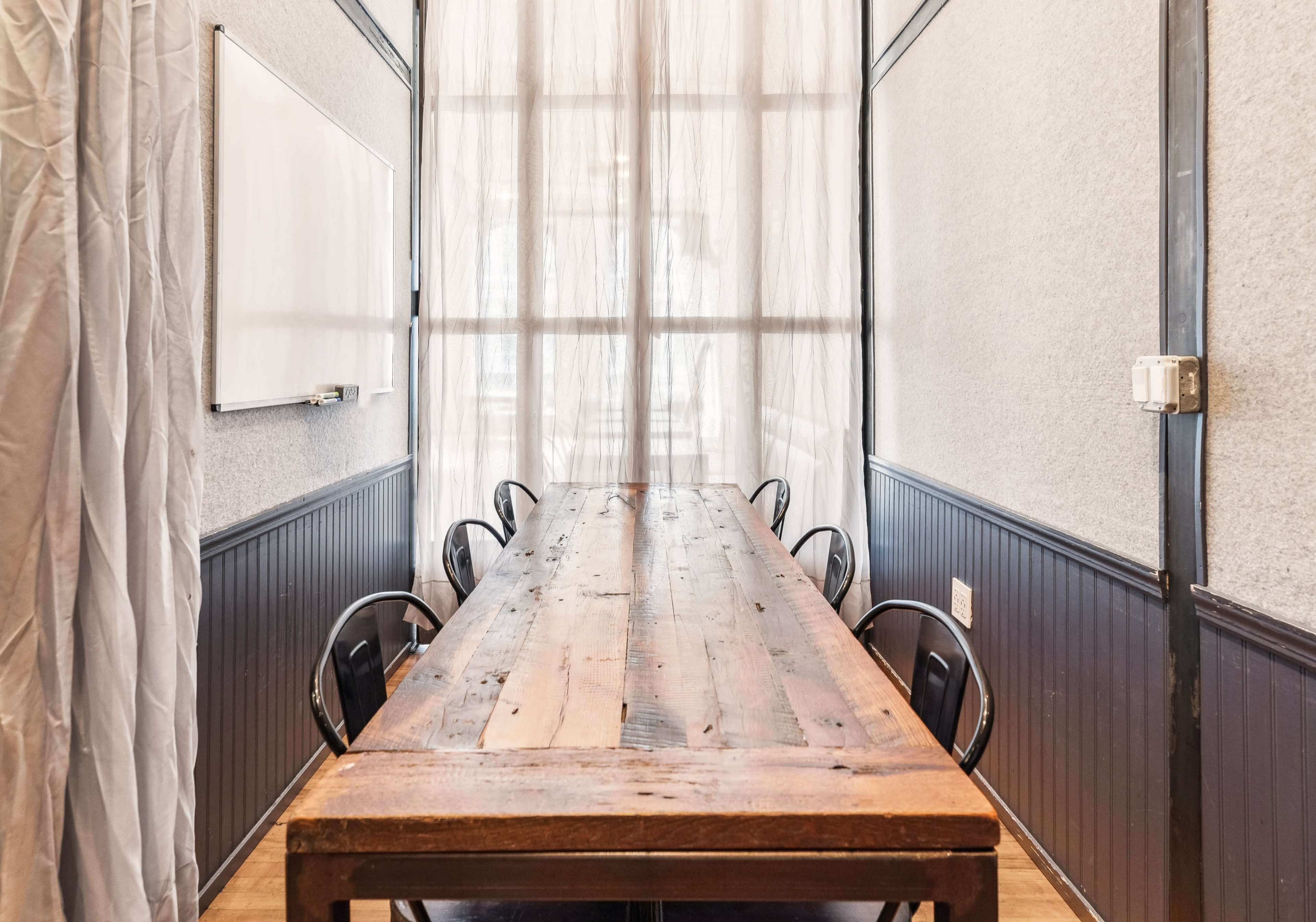 A long wooden table with several black chairs is situated in a meeting room, featuring large windows covered by sheer curtains.