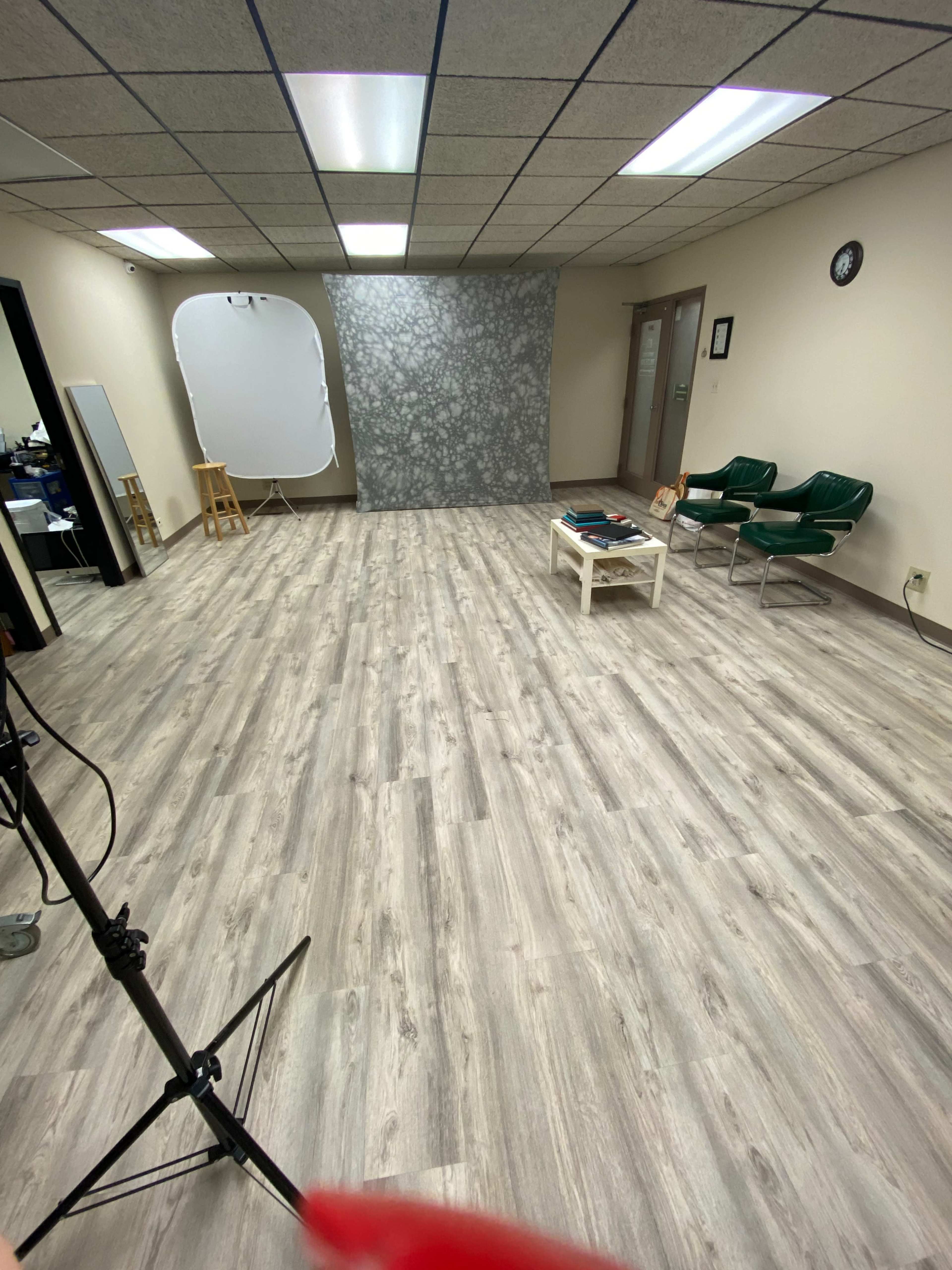 A spacious interior room with light wood flooring, a gray patterned backdrop, two green chairs, and a small table, set up for a photo session.