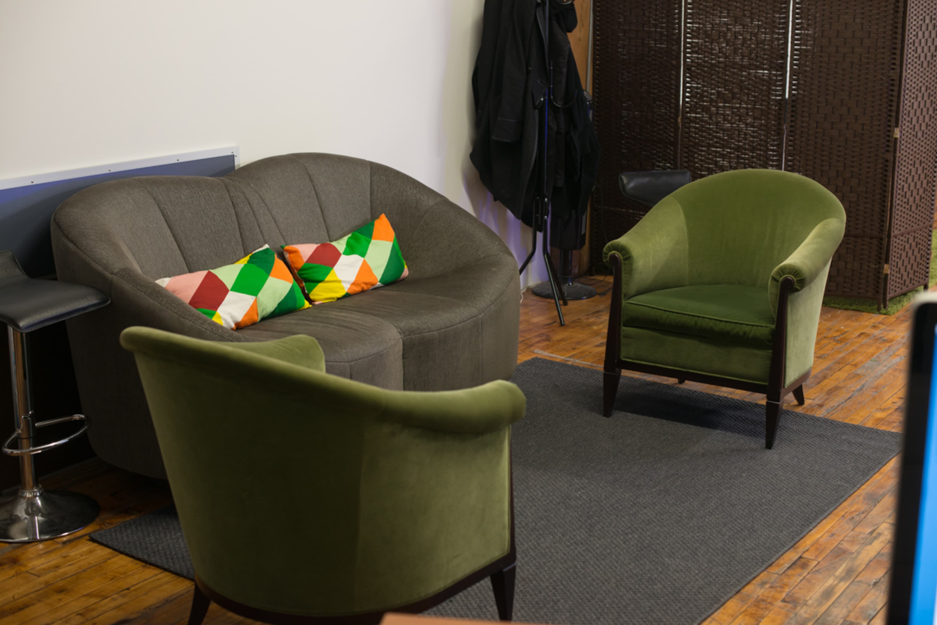 The image shows a cozy seating area featuring a curved gray sofa with colorful pillows, two green upholstered chairs, and a patterned rug on a wooden floor.