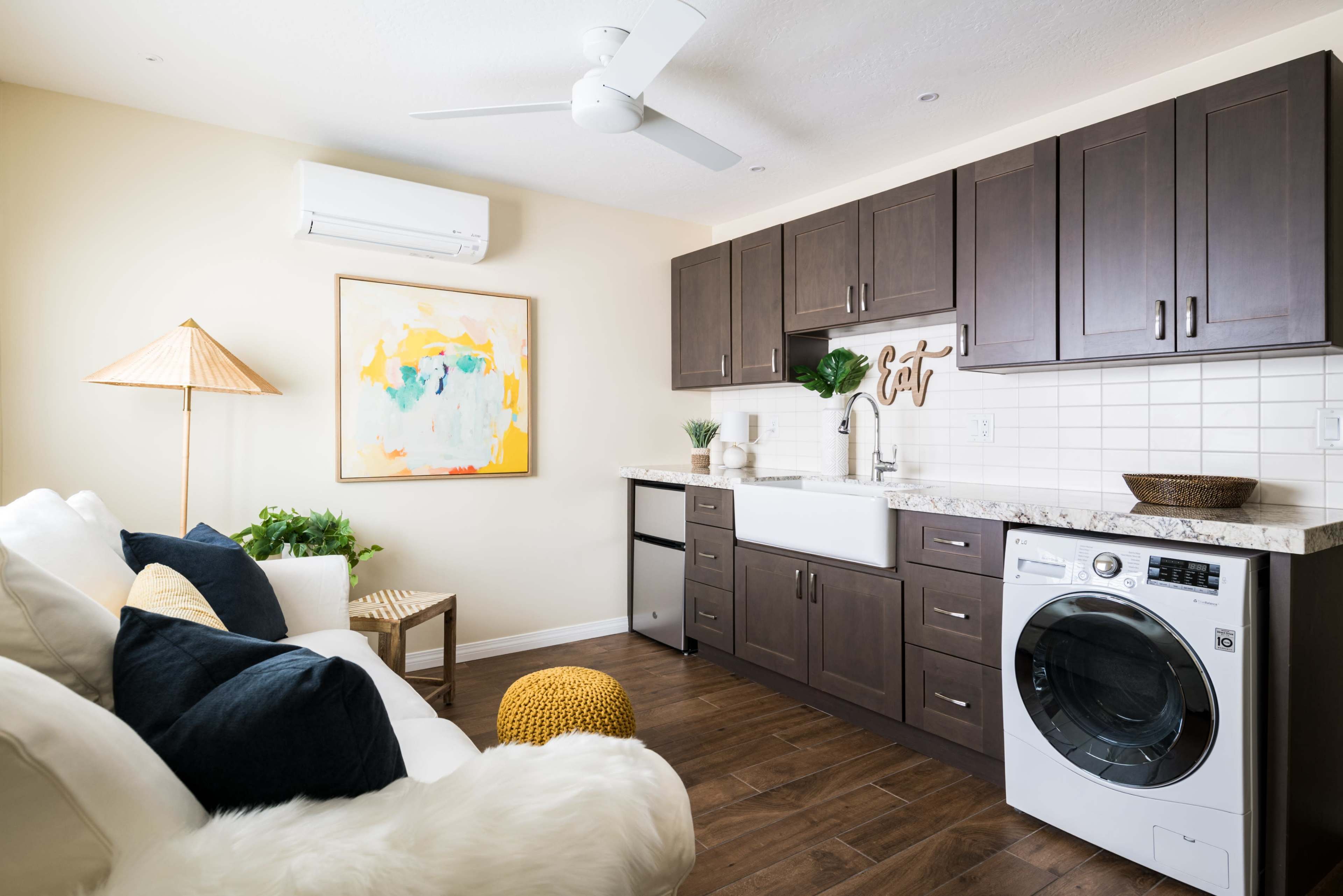 The image shows a modern laundry room featuring dark wooden cabinetry, a large sink, a washing machine, and decorative elements like plants and artwork.