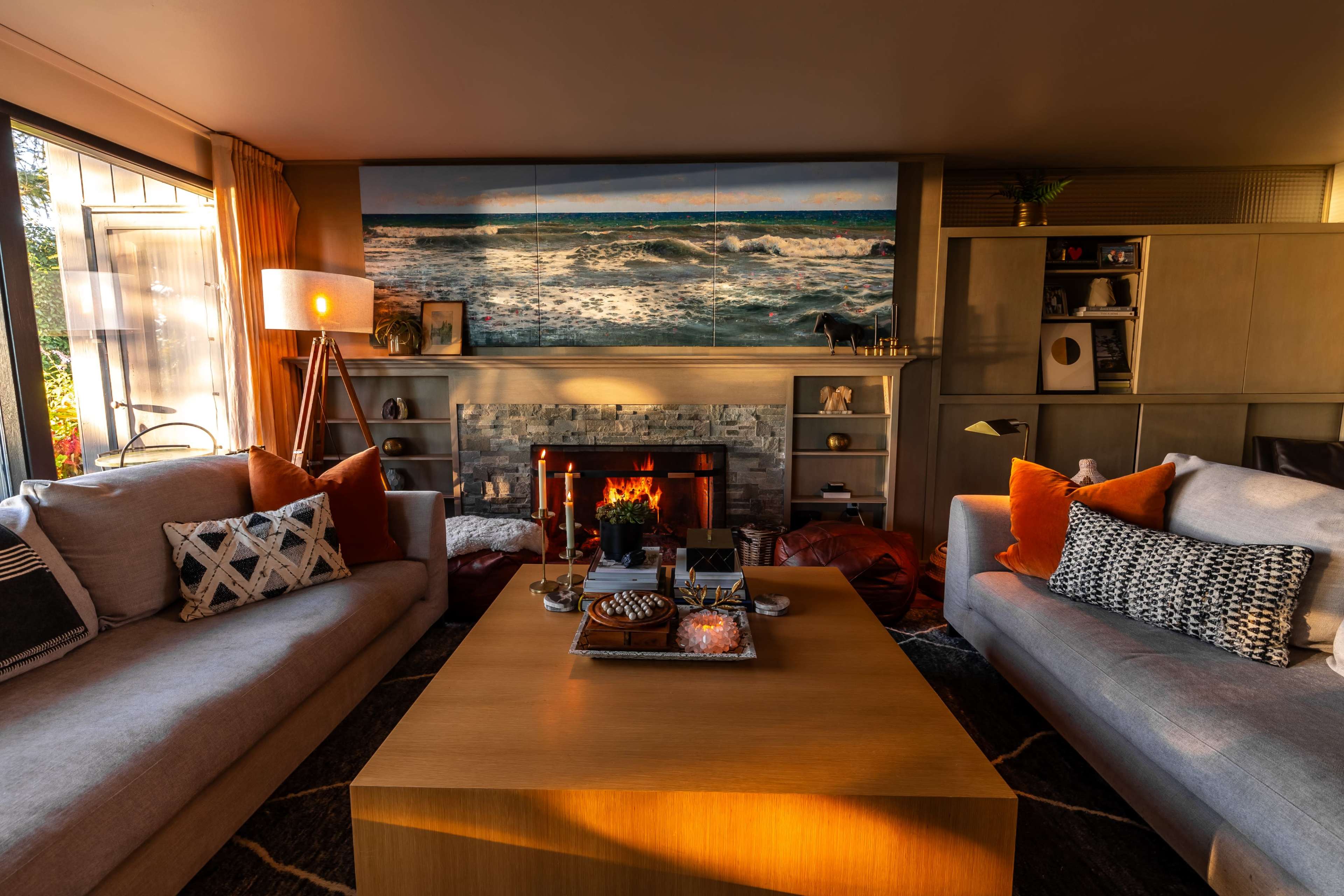 A cozy living room with two sofas facing a coffee table, a fireplace with a flame, and artwork of ocean waves on the wall.