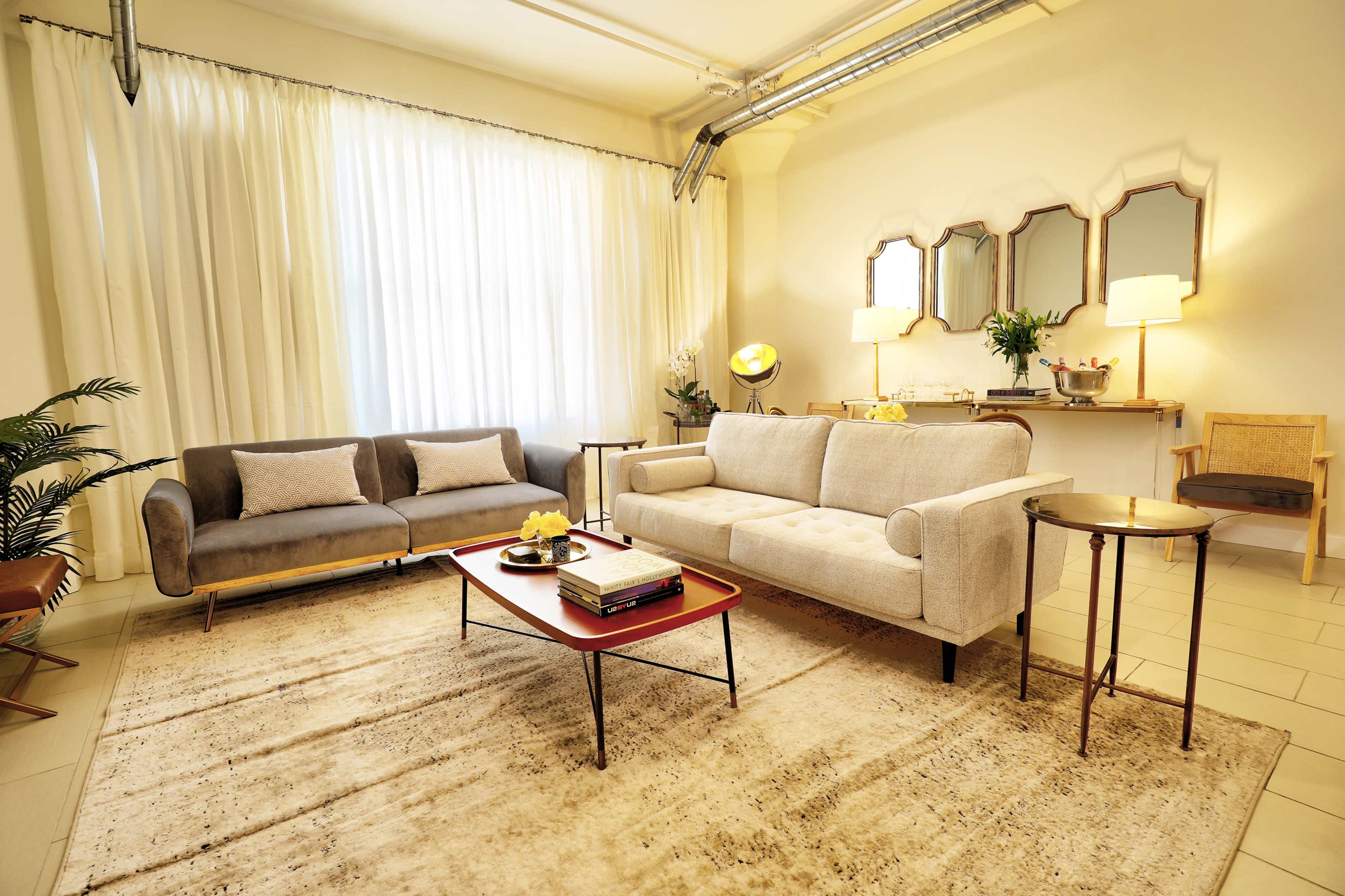 The image depicts a modern living room with a light-colored couch, a gray sofa, a coffee table, and a side table, all arranged on a soft area rug beneath large windows with sheer curtains.