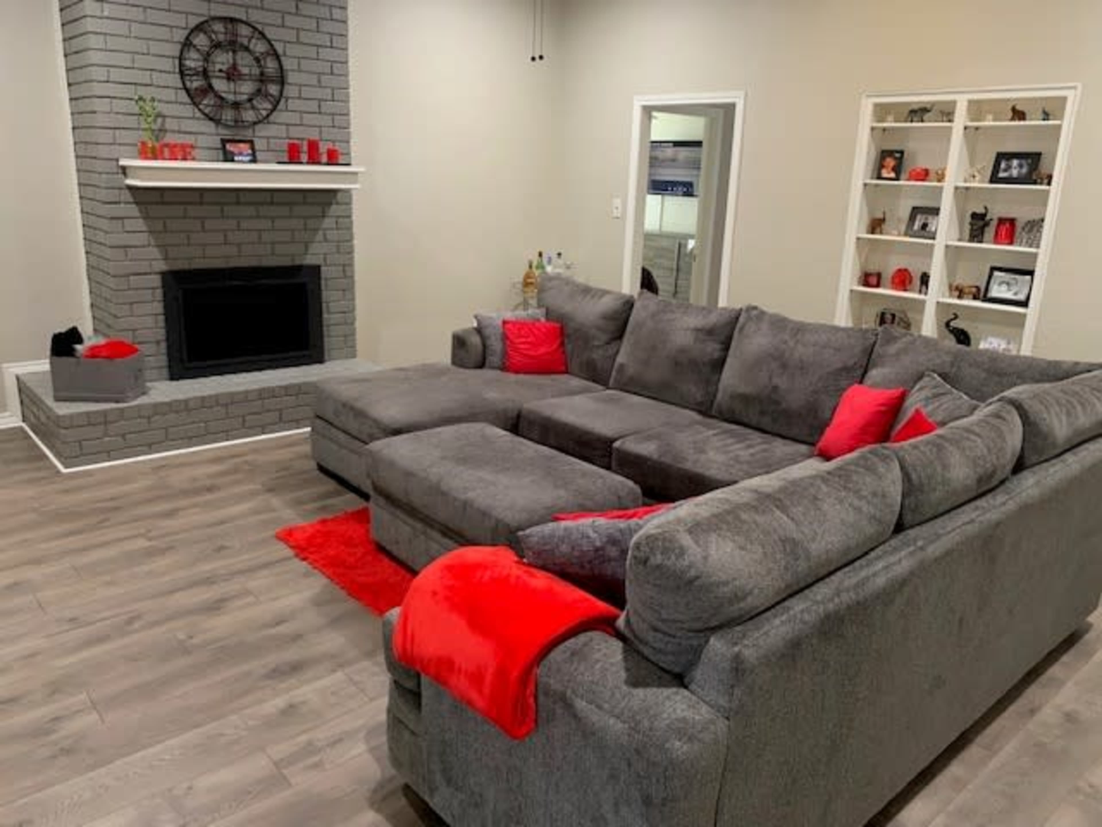 A spacious living room featuring a gray sectional sofa with red accent pillows, a brick fireplace, and shelves displaying framed photos and decorative items.