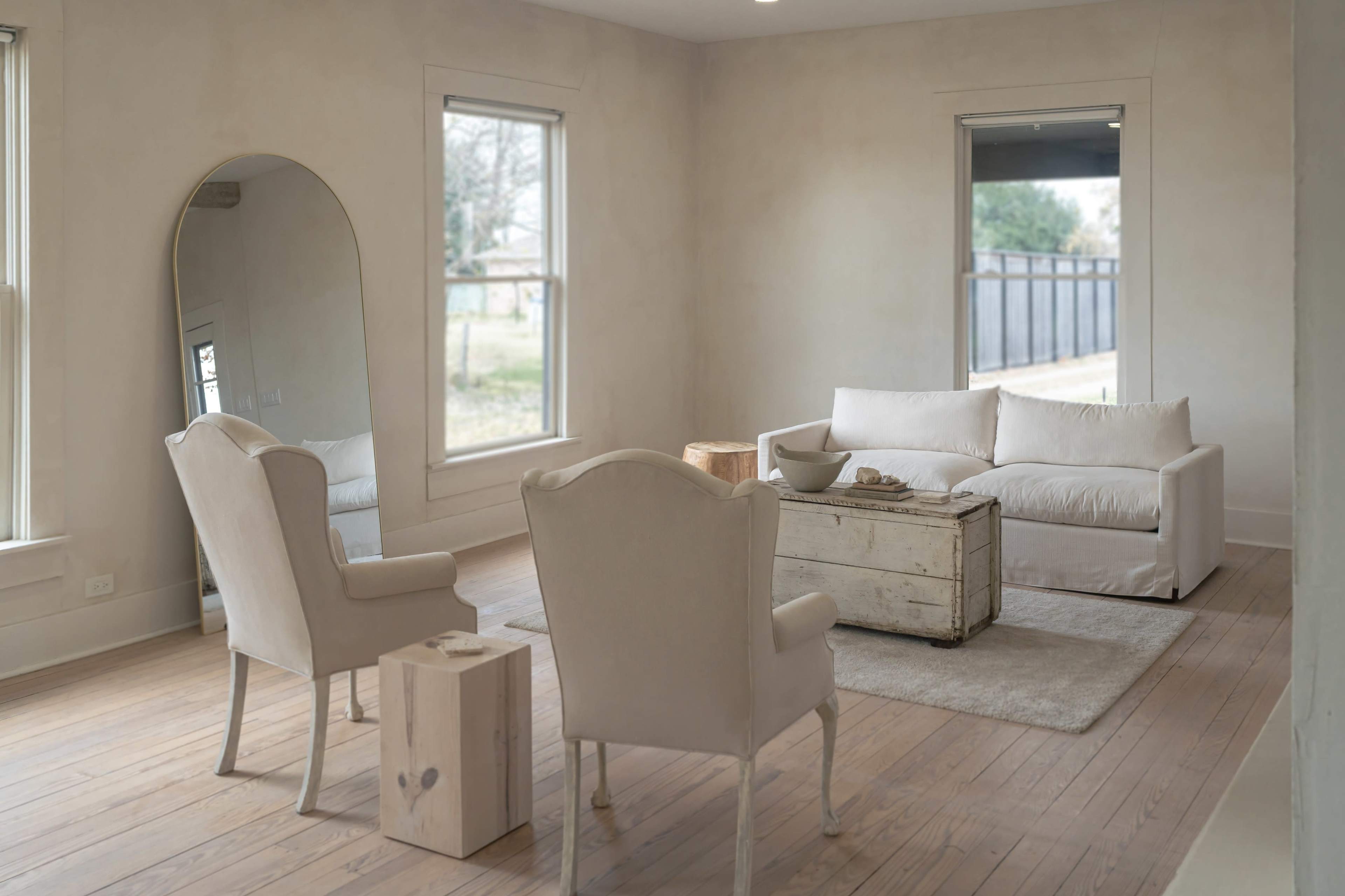 The image shows a minimalist living room featuring a white sofa, two matching armchairs, a wooden coffee table, and large windows letting in natural light.