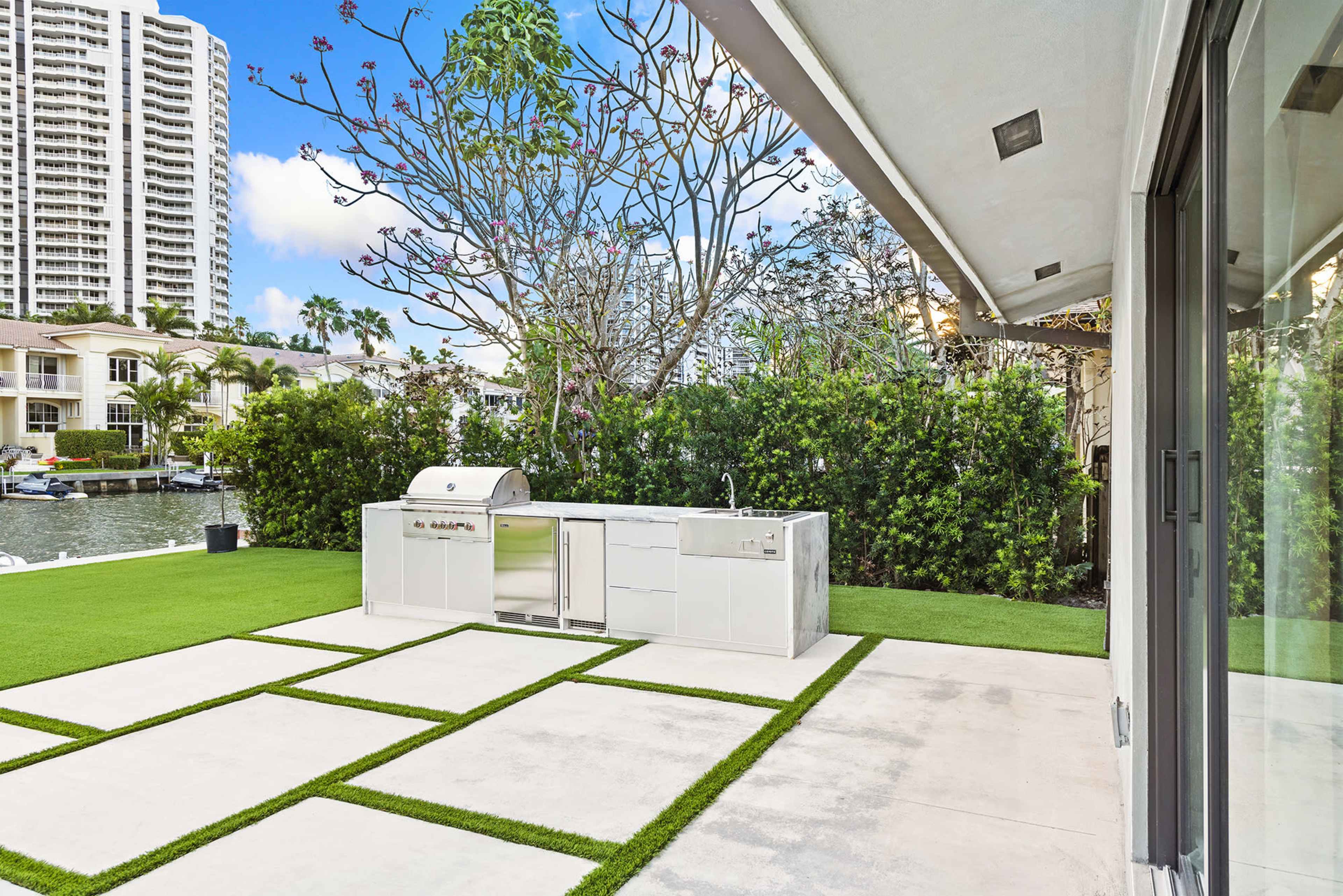 An outdoor kitchen area with a stainless steel grill, situated on a landscaped patio with rectangular concrete slabs and lush green grass, overlooking a waterfront view.