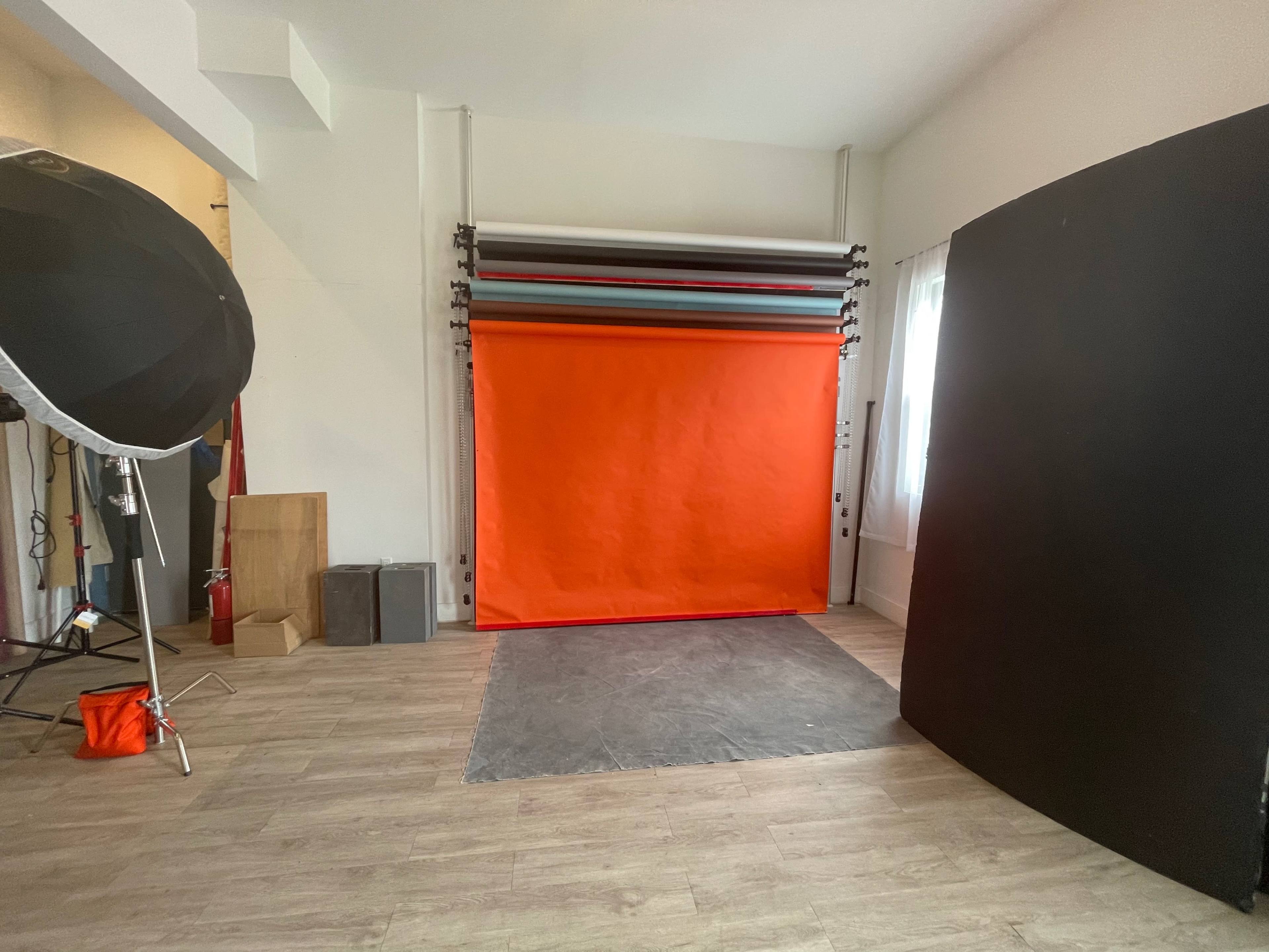 A photography studio with an orange backdrop, various lighting equipment, and a dark panel in a well-lit space.