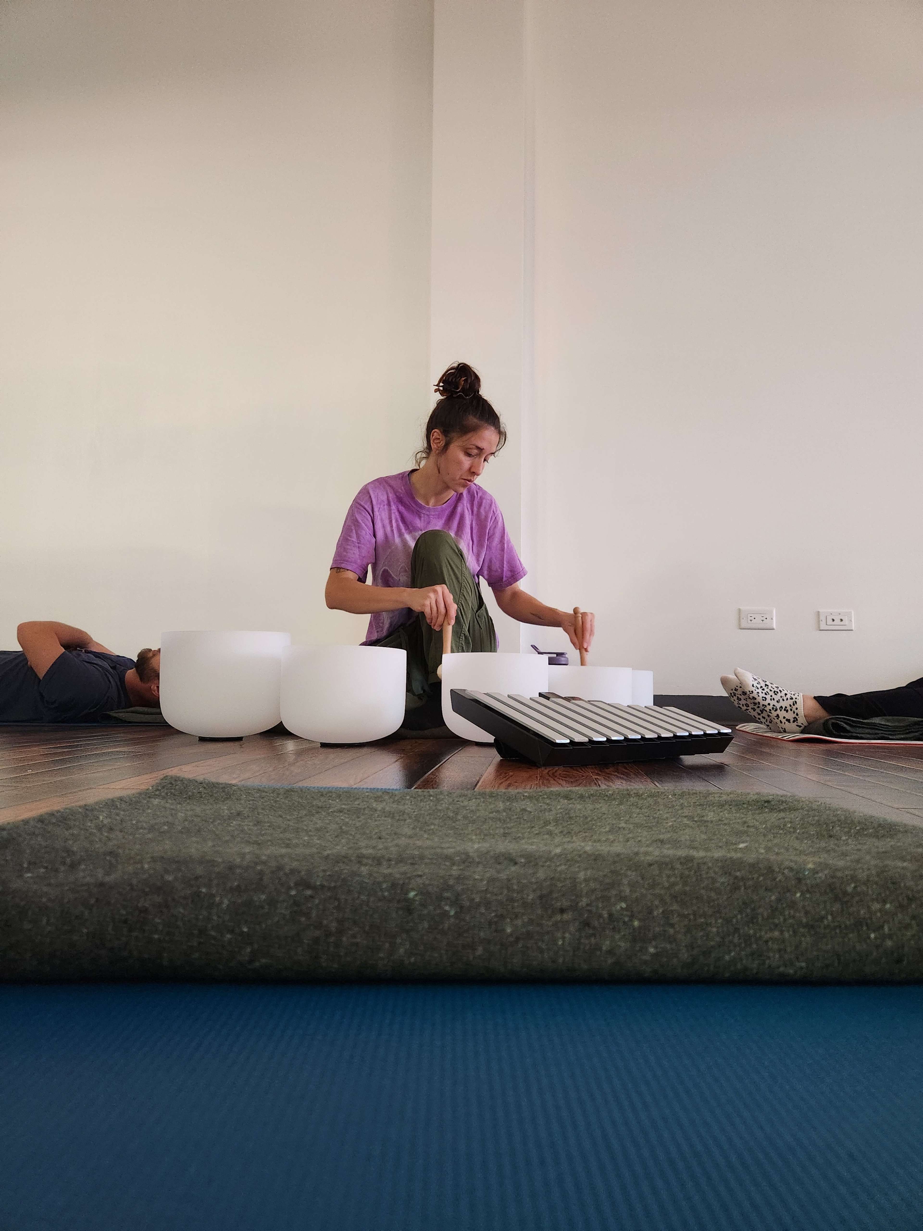 A woman in a purple shirt plays a musical instrument while sitting on the floor, surrounded by two sound bowls and several participants lying on yoga mats.