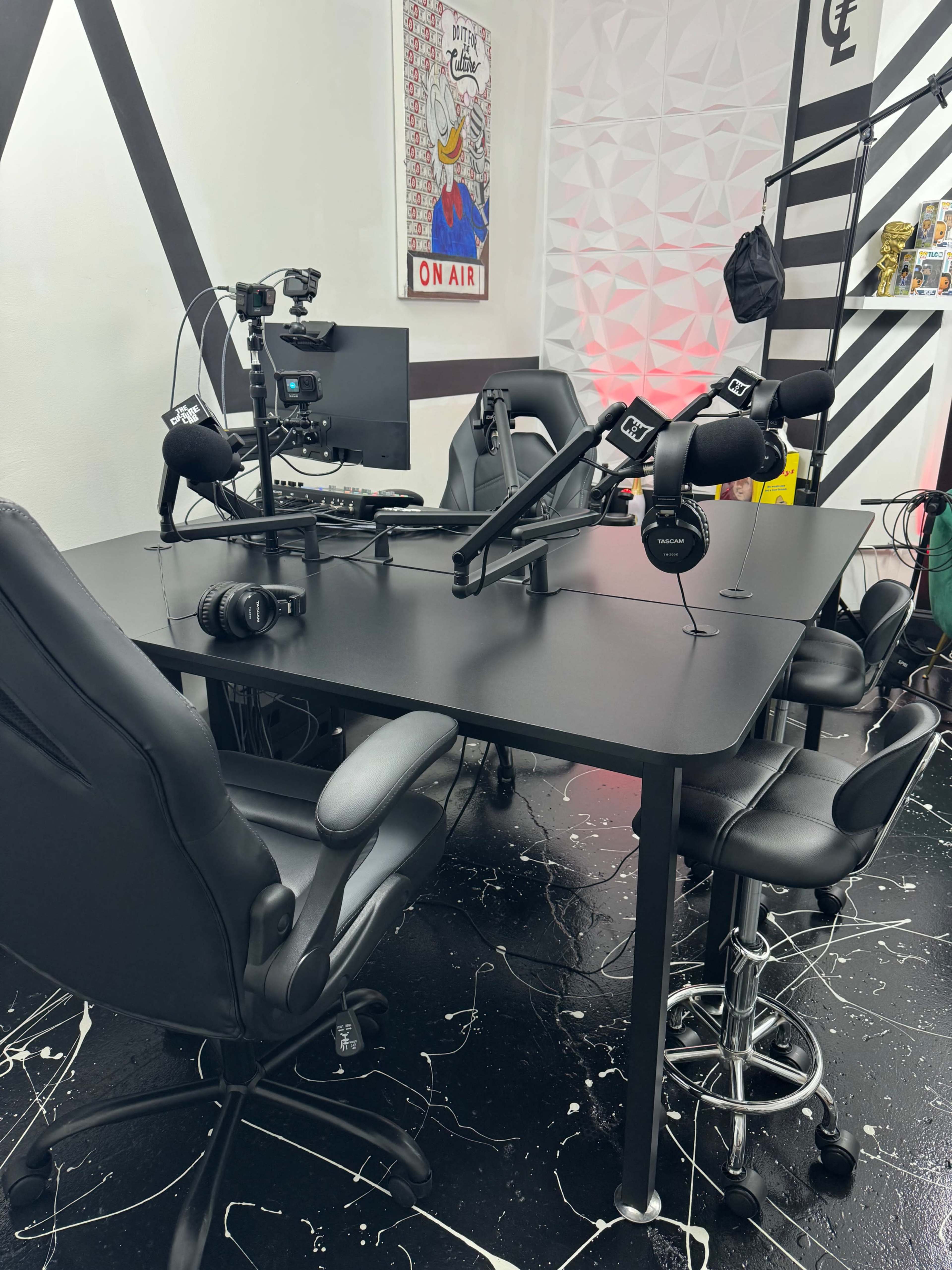 A black podcast studio setup featuring a table, four chairs, microphones on adjustable arms, a computer monitor, and headphones.