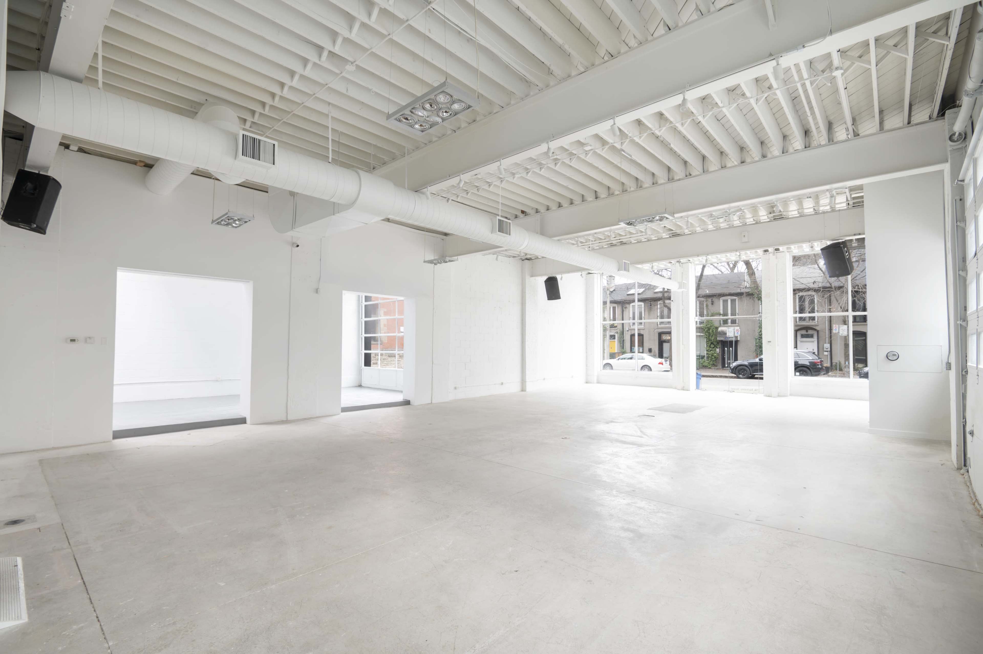 The image shows a large, empty interior space with white walls and a concrete floor, featuring exposed ductwork and large windows letting in natural light.