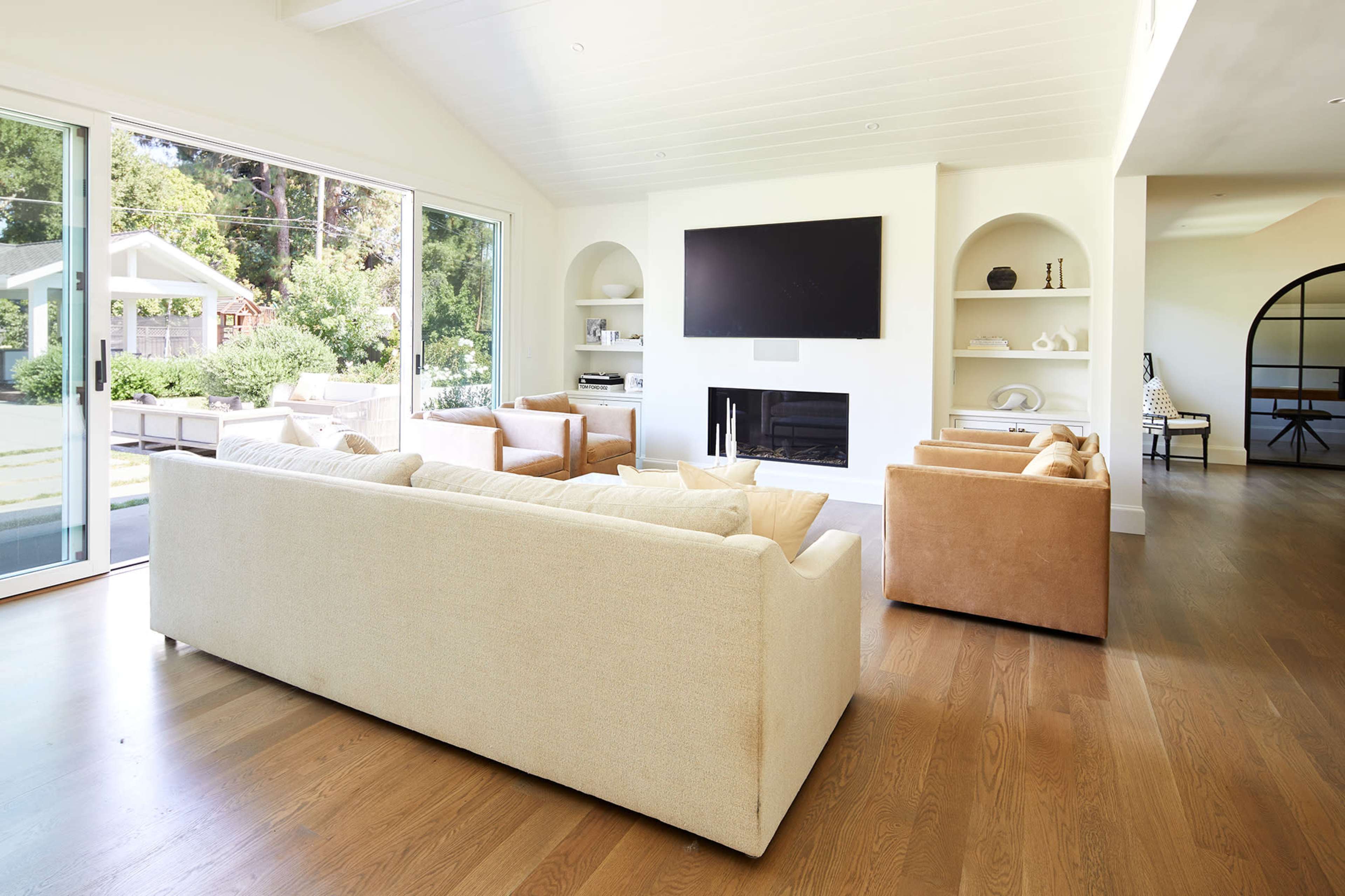A modern living room features a light-colored sofa and armchairs arranged around a wall-mounted television and a fireplace, with large windows overlooking a green outdoor space.