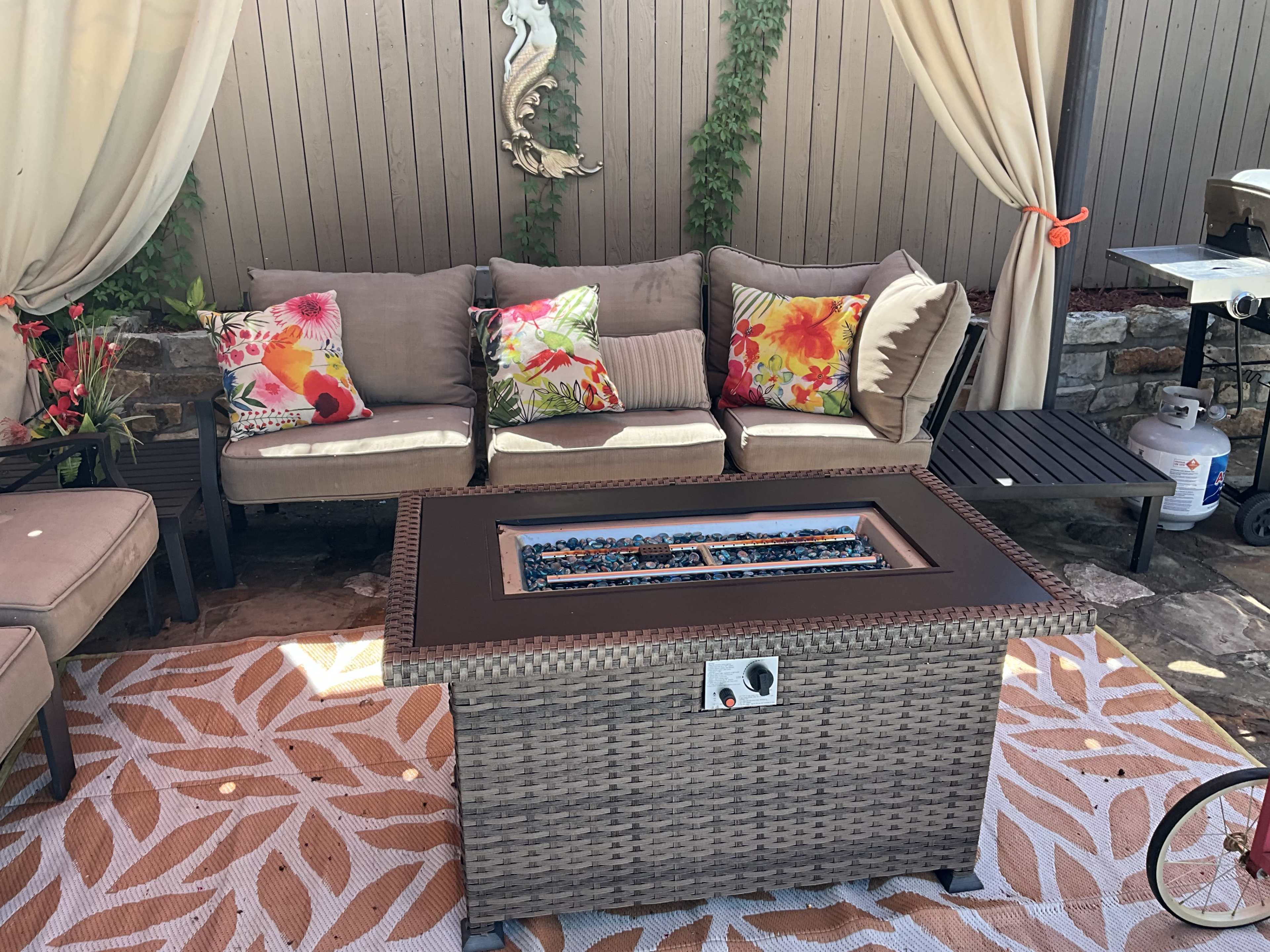 A patio area with a woven coffee table featuring a glass-top fire pit surrounded by a sectional sofa with floral-patterned cushions and draped curtains.