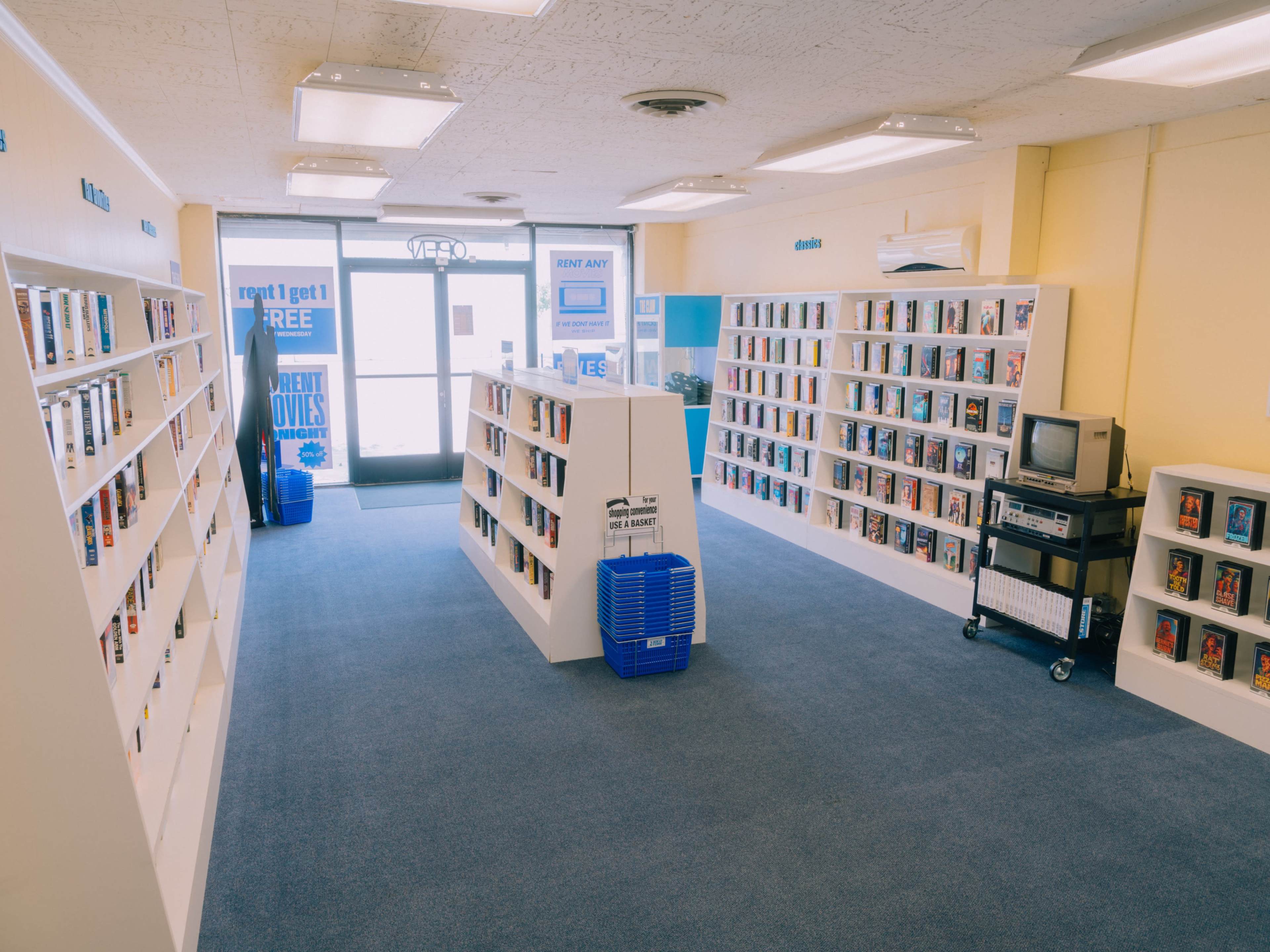 The image shows an empty video rental store with shelves lined with DVDs and a small television display in the center.