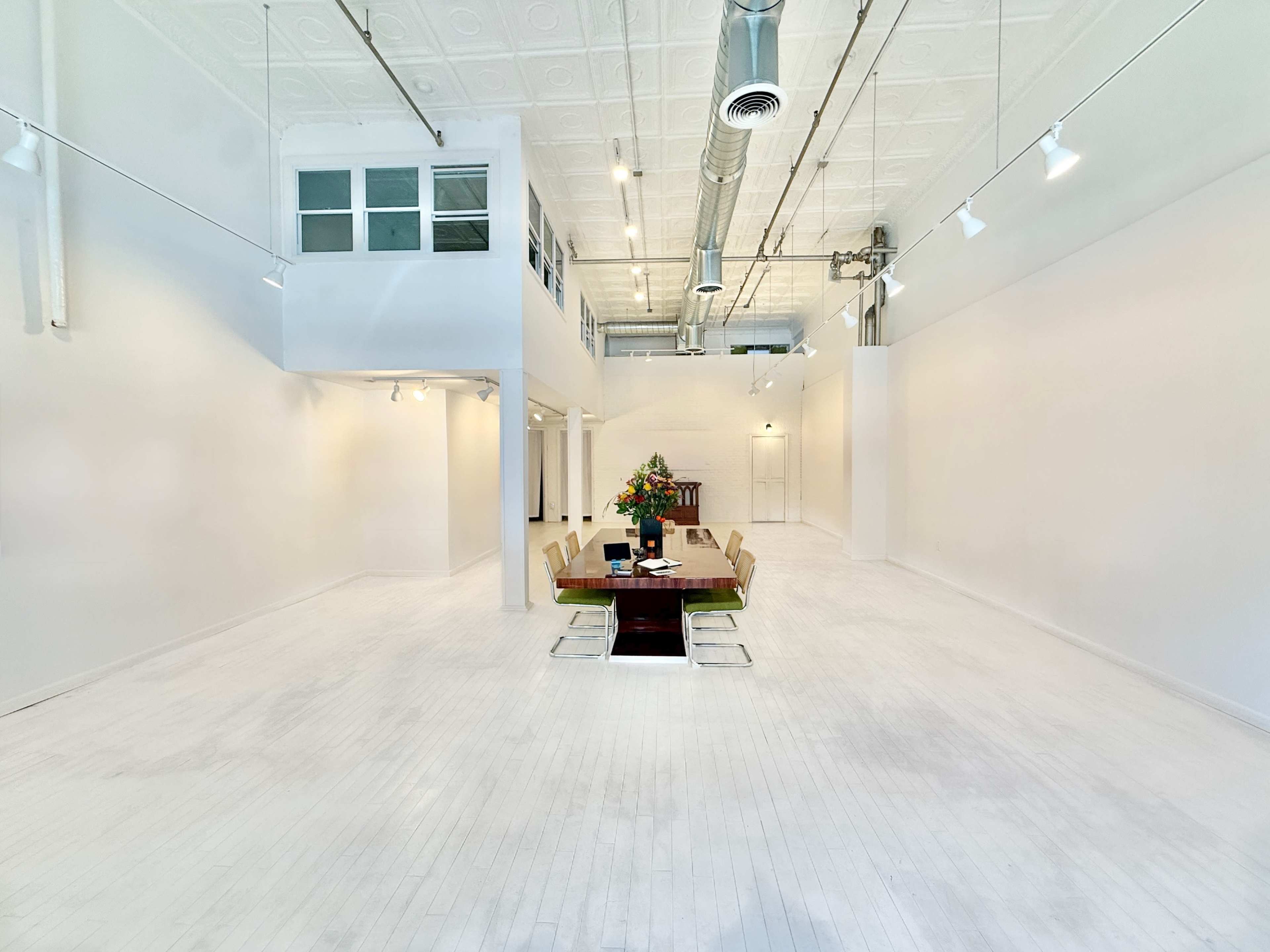 The image shows a spacious, brightly lit room with white walls and flooring, featuring a long dining table set with chairs and a floral arrangement.