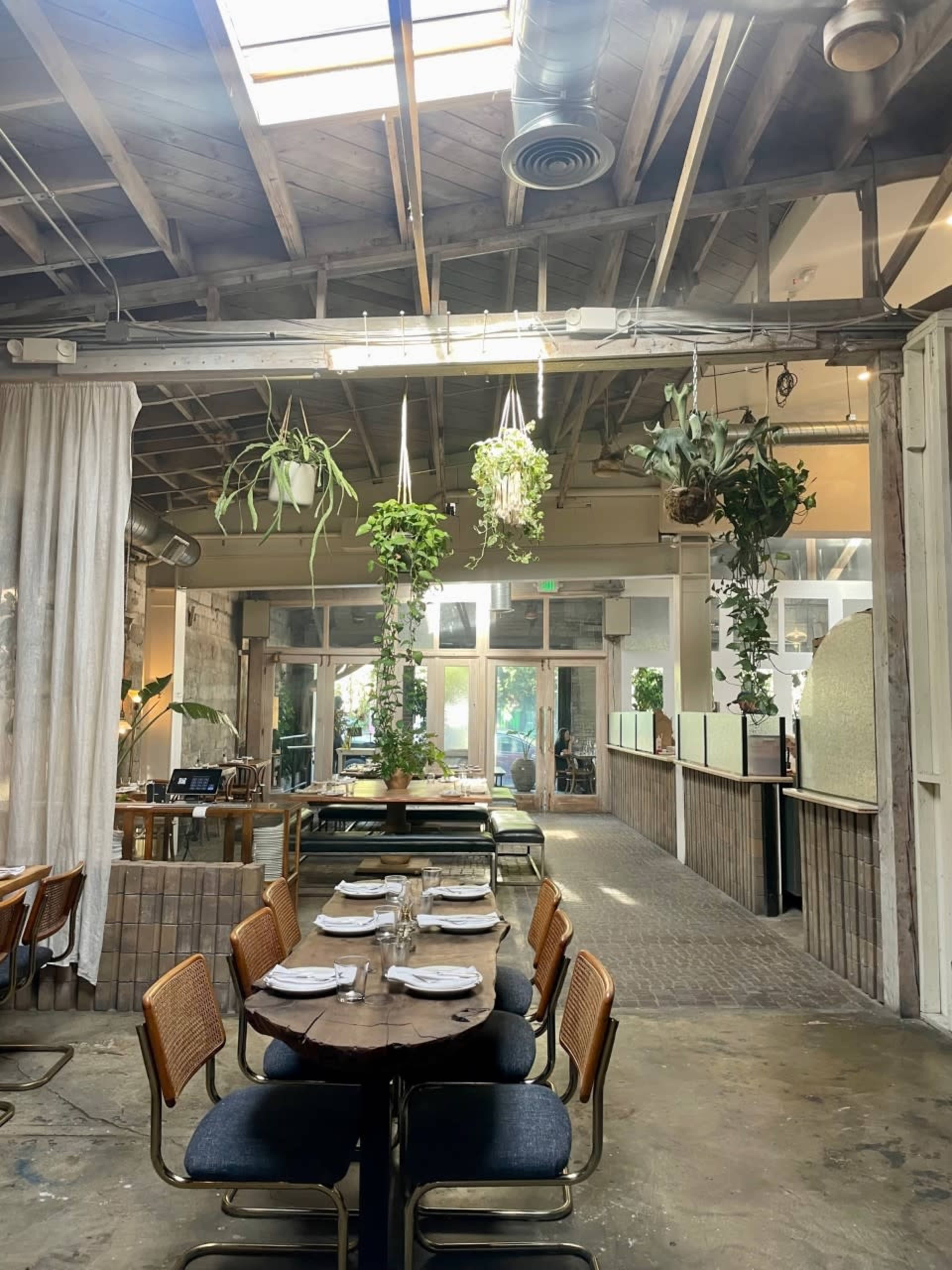 A modern restaurant interior featuring a long wooden table set for dining, with hanging plants and large windows allowing natural light to enter the space.