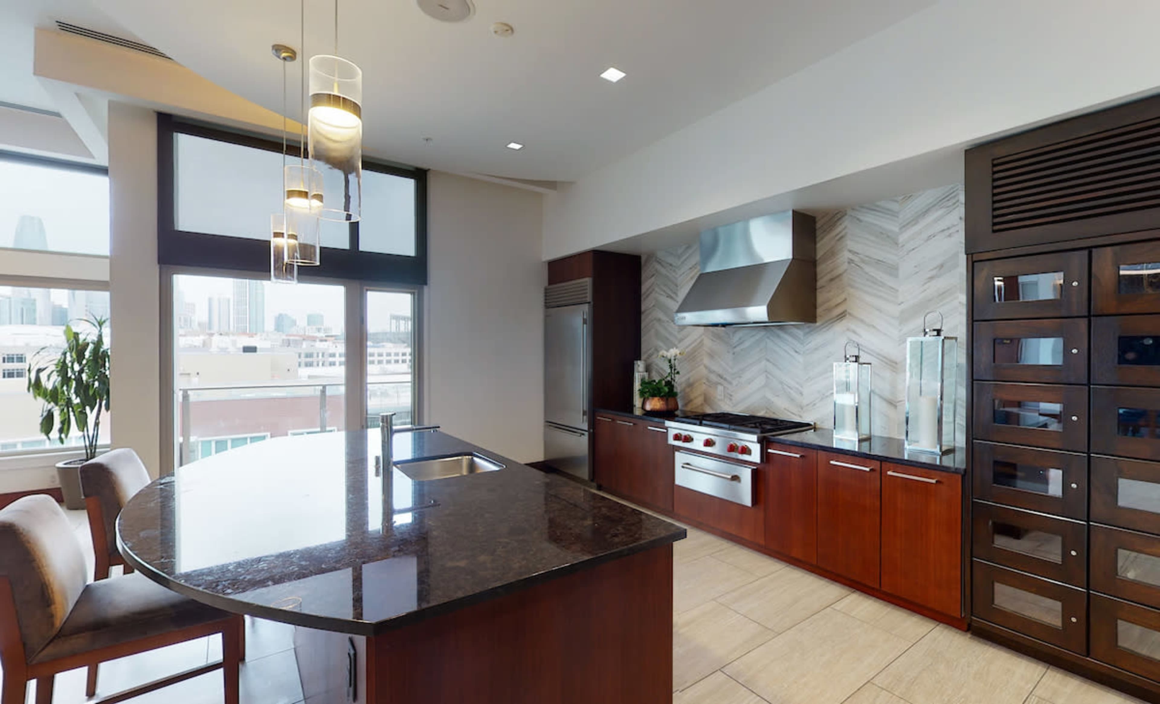 A modern kitchen features a large island, stainless steel appliances, and a tiled backsplash with city views through large windows.