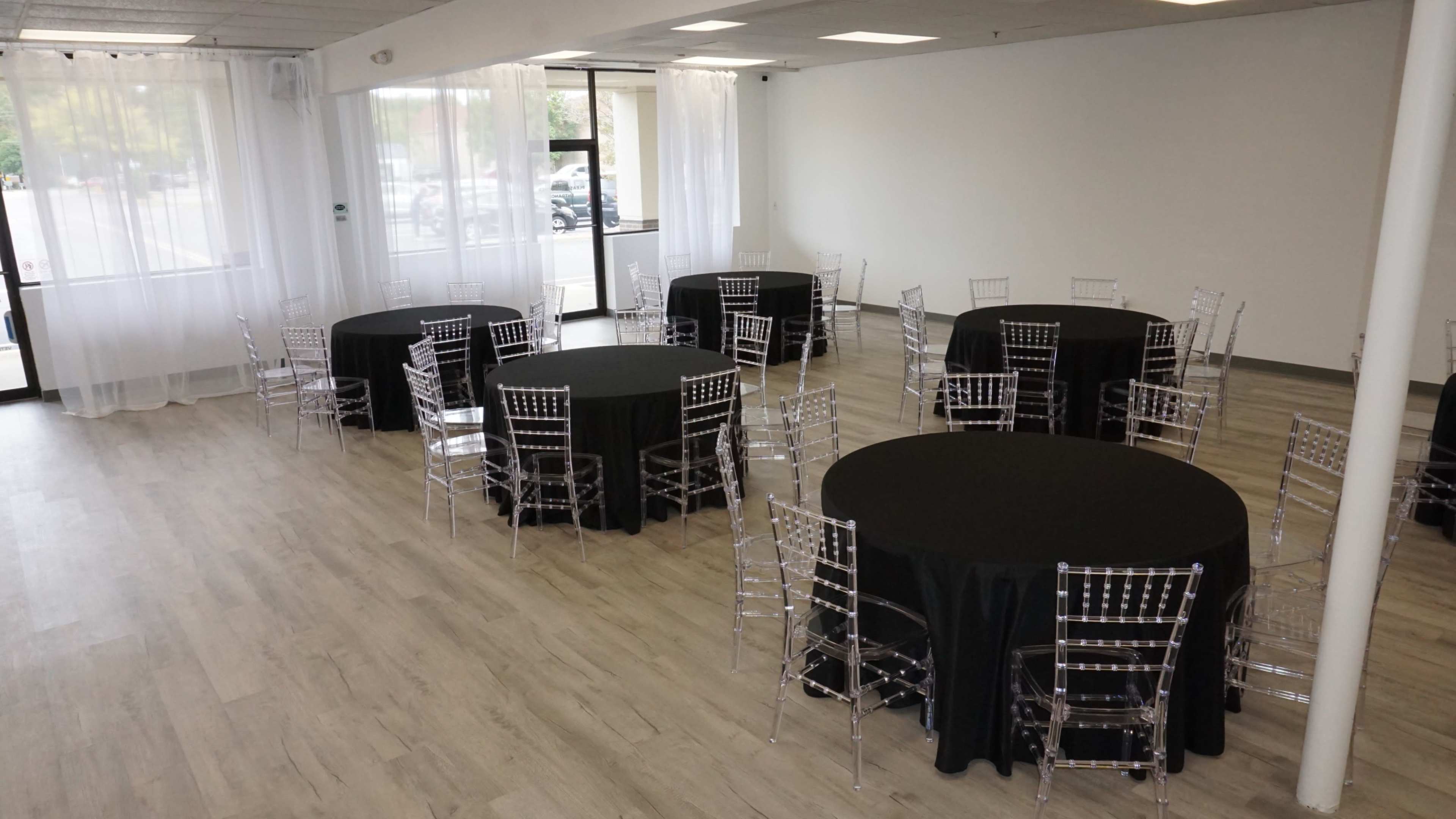 The image shows a spacious event room with several round tables covered in black tablecloths and surrounded by clear acrylic chairs.