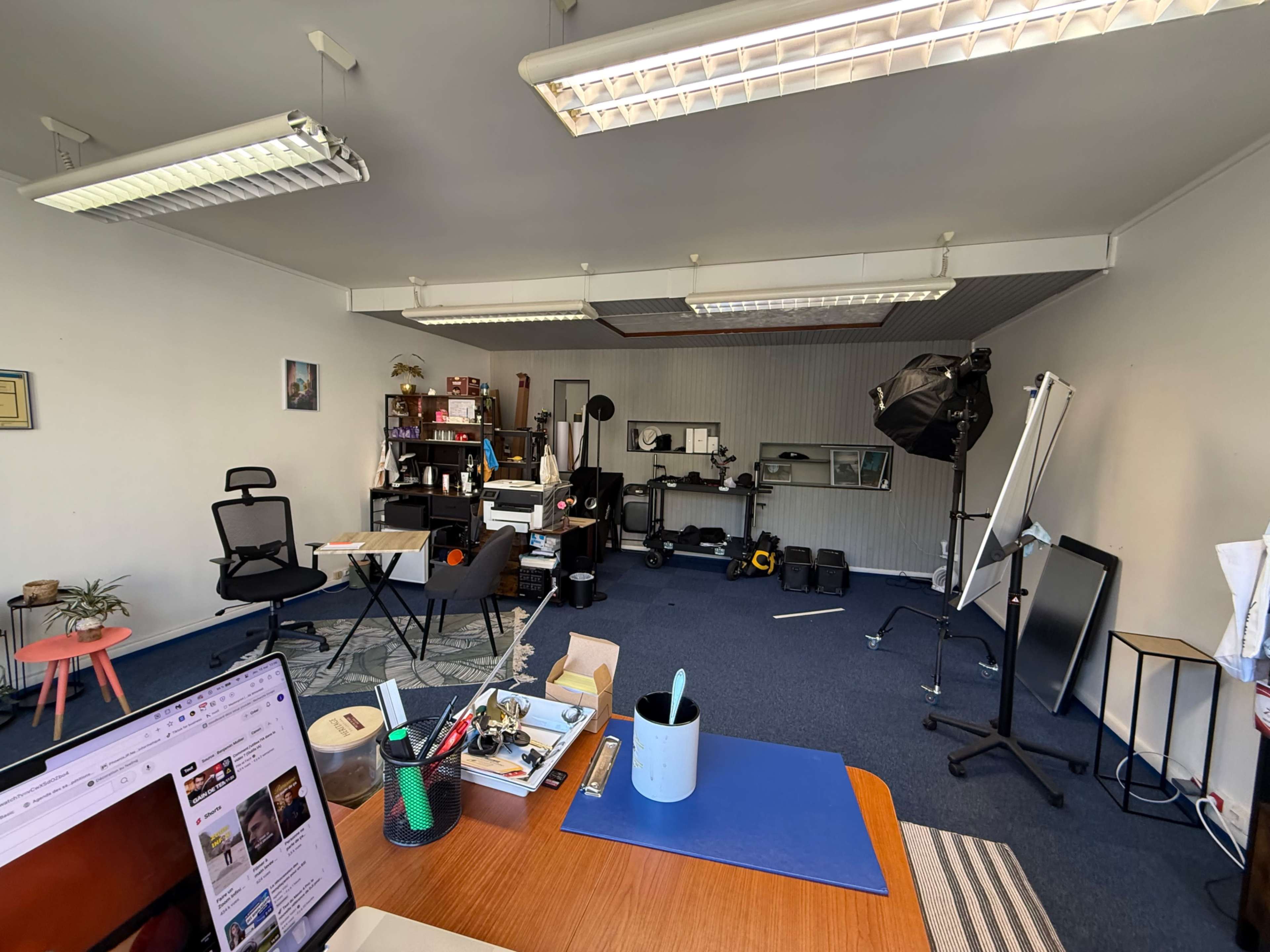 The image shows a spacious workspace with various office furniture and equipment, including a desk, chairs, and photography gear.