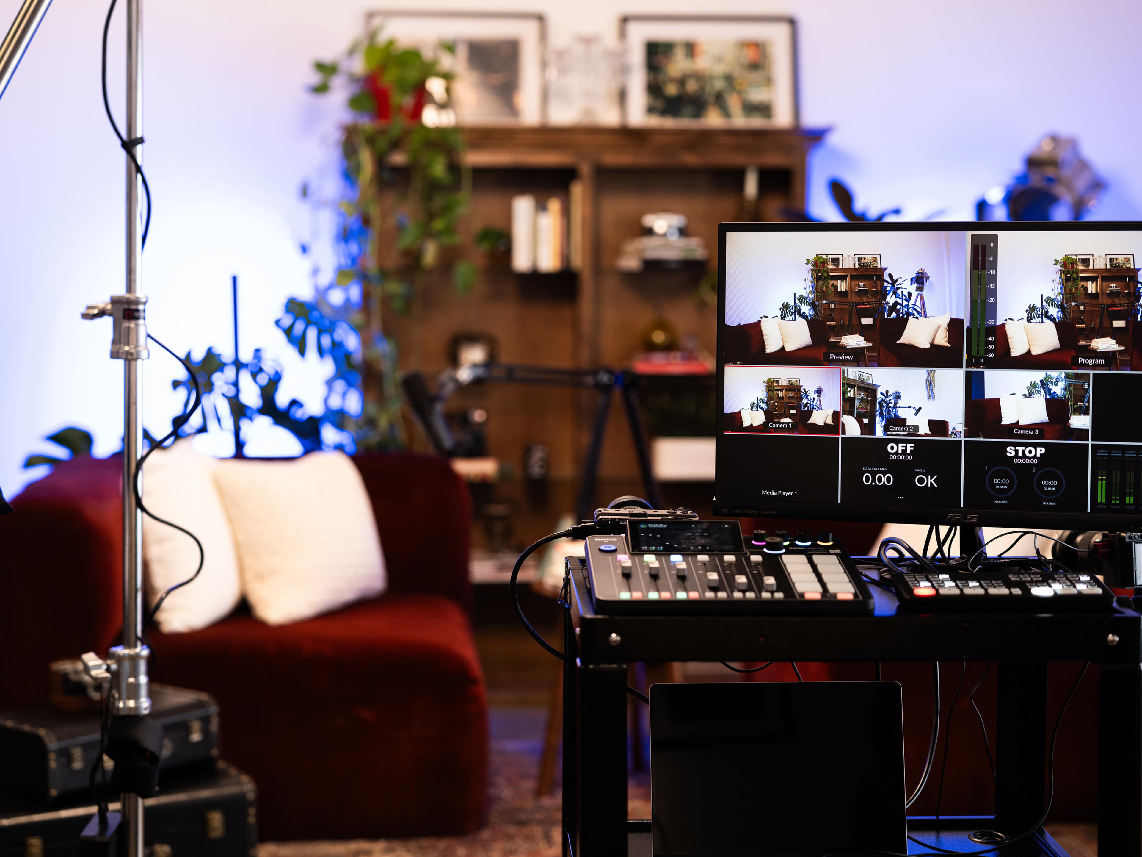 A recording setup with multiple screens displaying a cozy room, featuring a red couch, a bookshelf, and various plants.