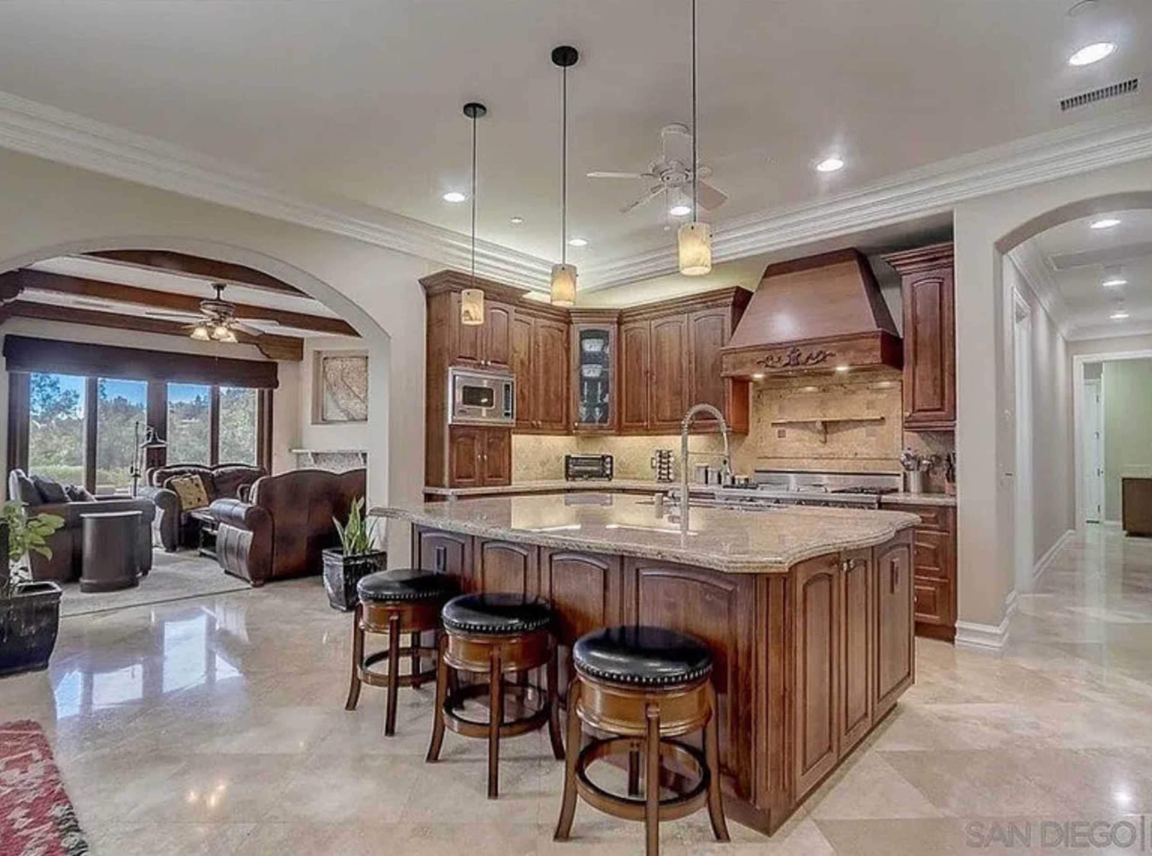 The image shows a spacious kitchen with wooden cabinetry, a large island with bar stools, and an adjacent living area visible through an archway.