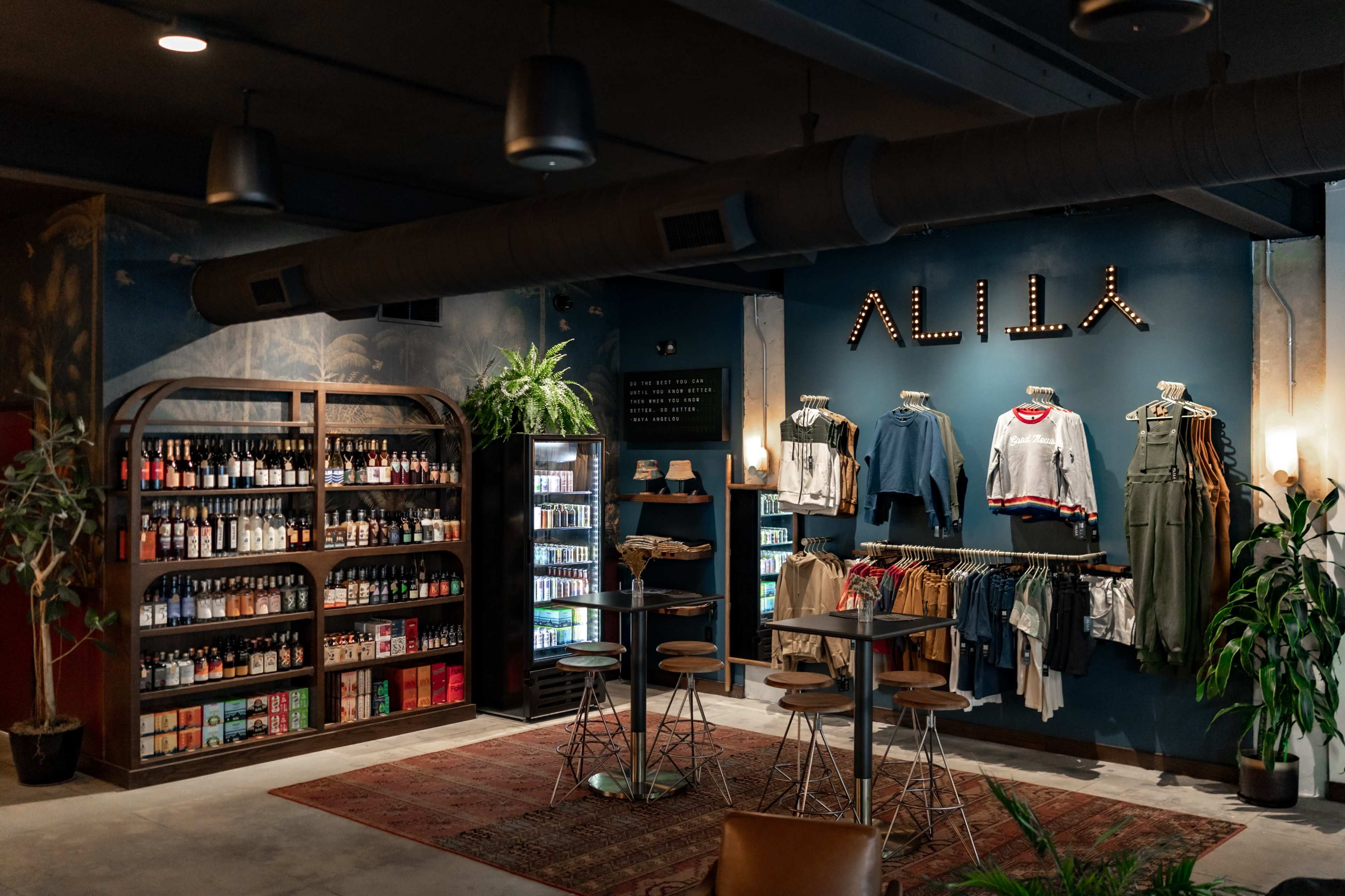 The image shows a stylish retail space featuring a display of clothing on racks, shelves of bottled beverages, and seating arrangements in a modern, well-lit ambiance.