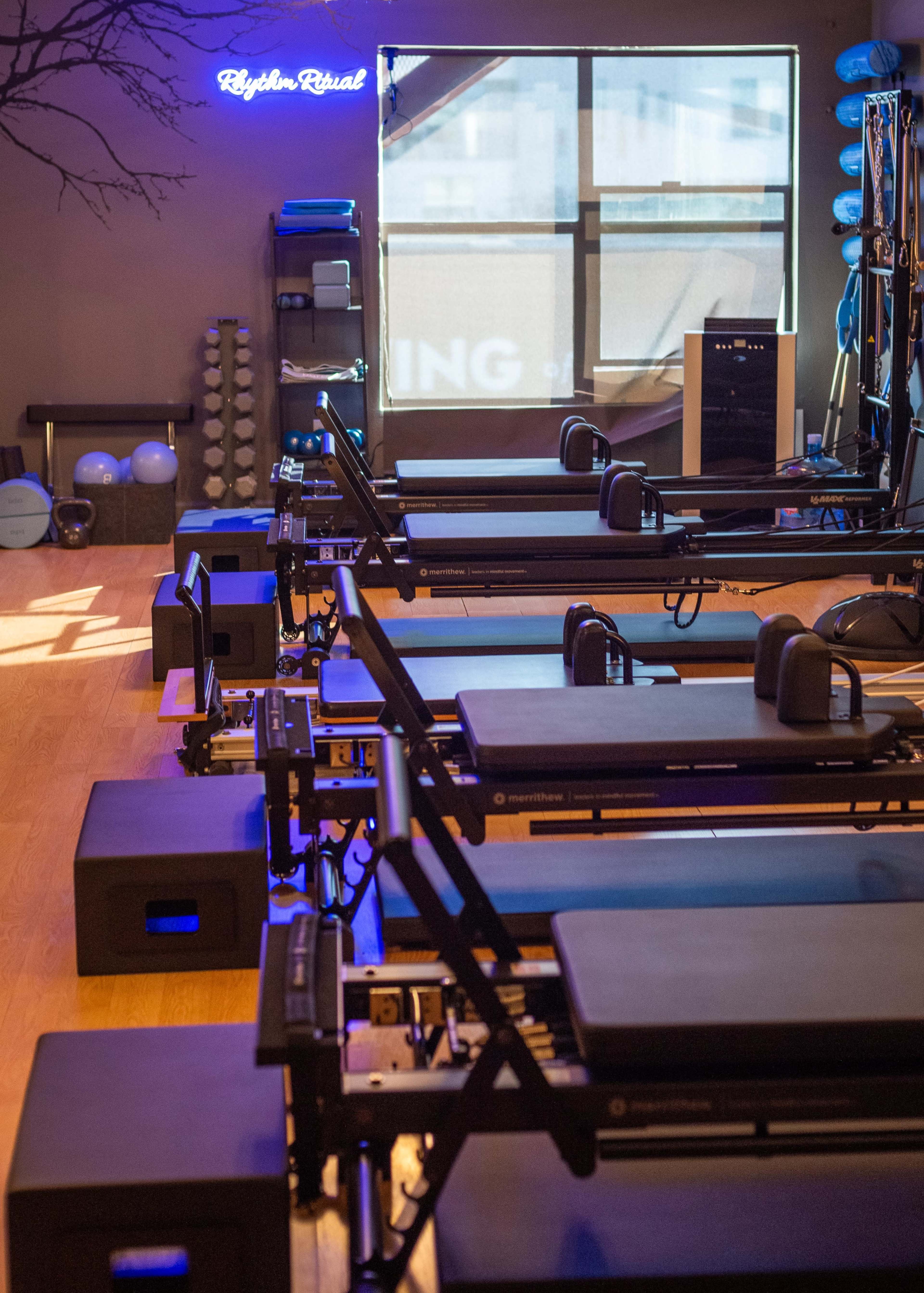 The image shows a fitness studio with several reformer machines arranged in a row, illuminated by soft lighting and featuring exercise equipment on the walls.