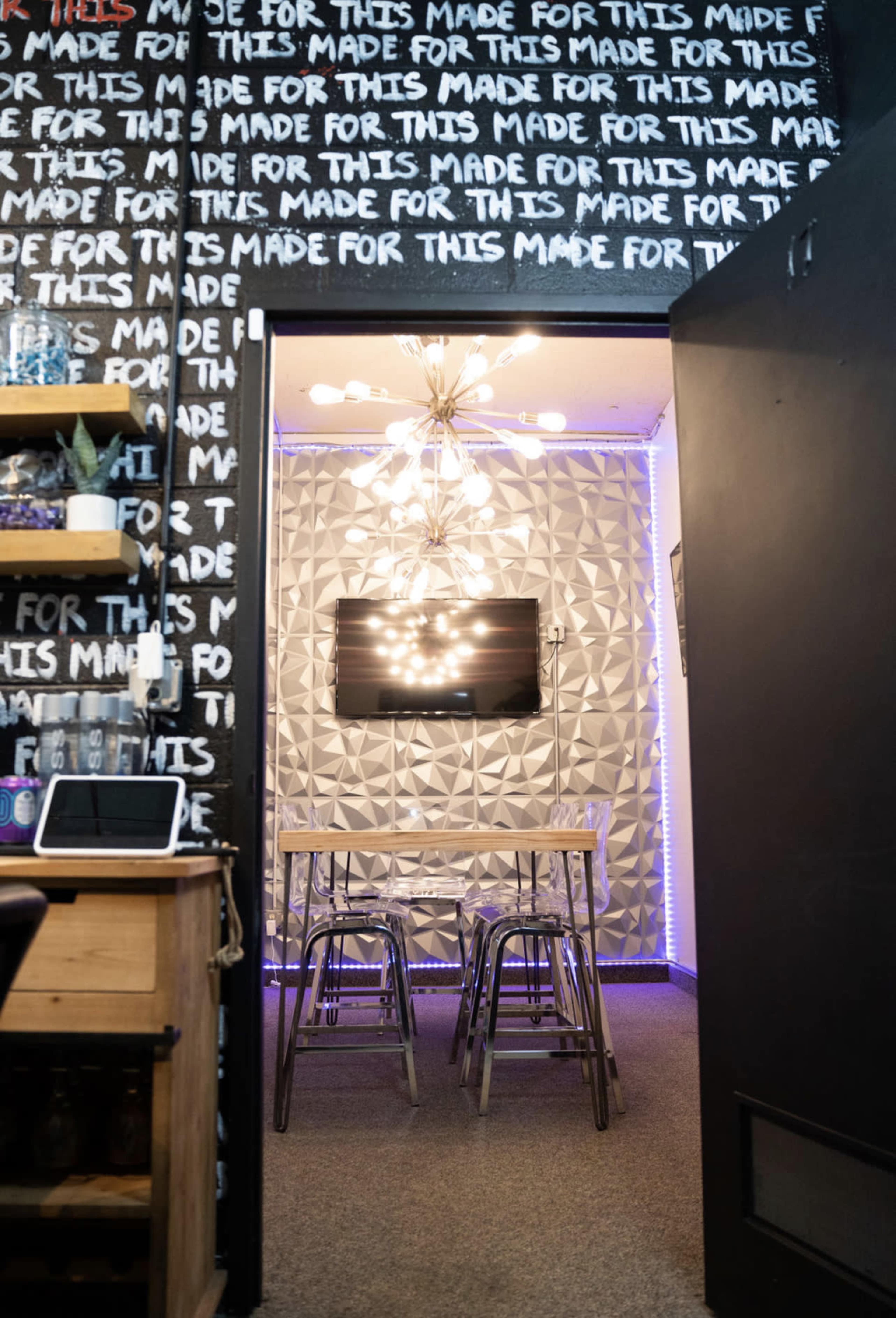 A modern room with a table and chairs, illuminated by a large chandelier, with a television mounted on a textured wall.