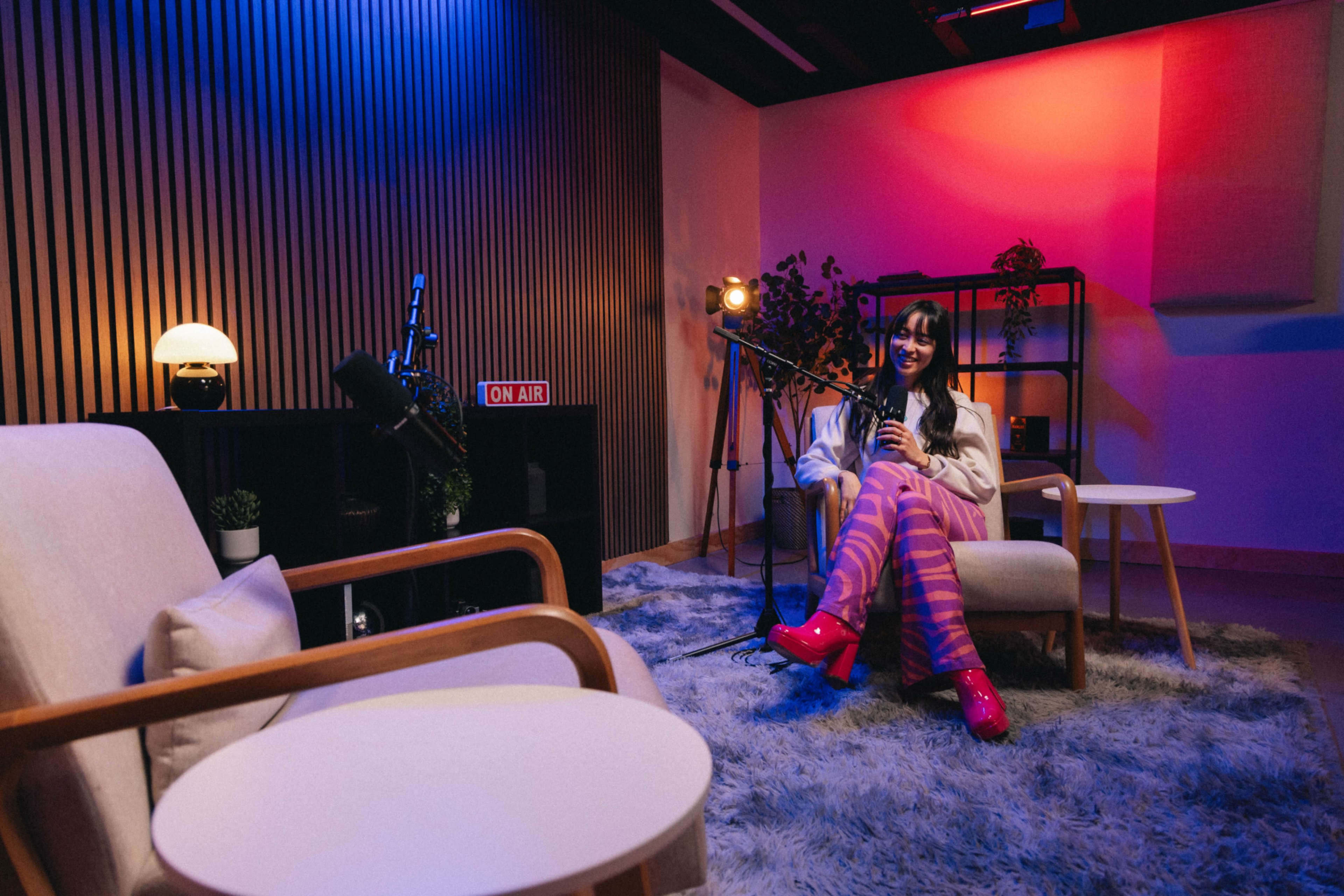 A young woman sits in a modern studio space with colorful lighting, holding a microphone, surrounded by cozy furniture and a recording setup.