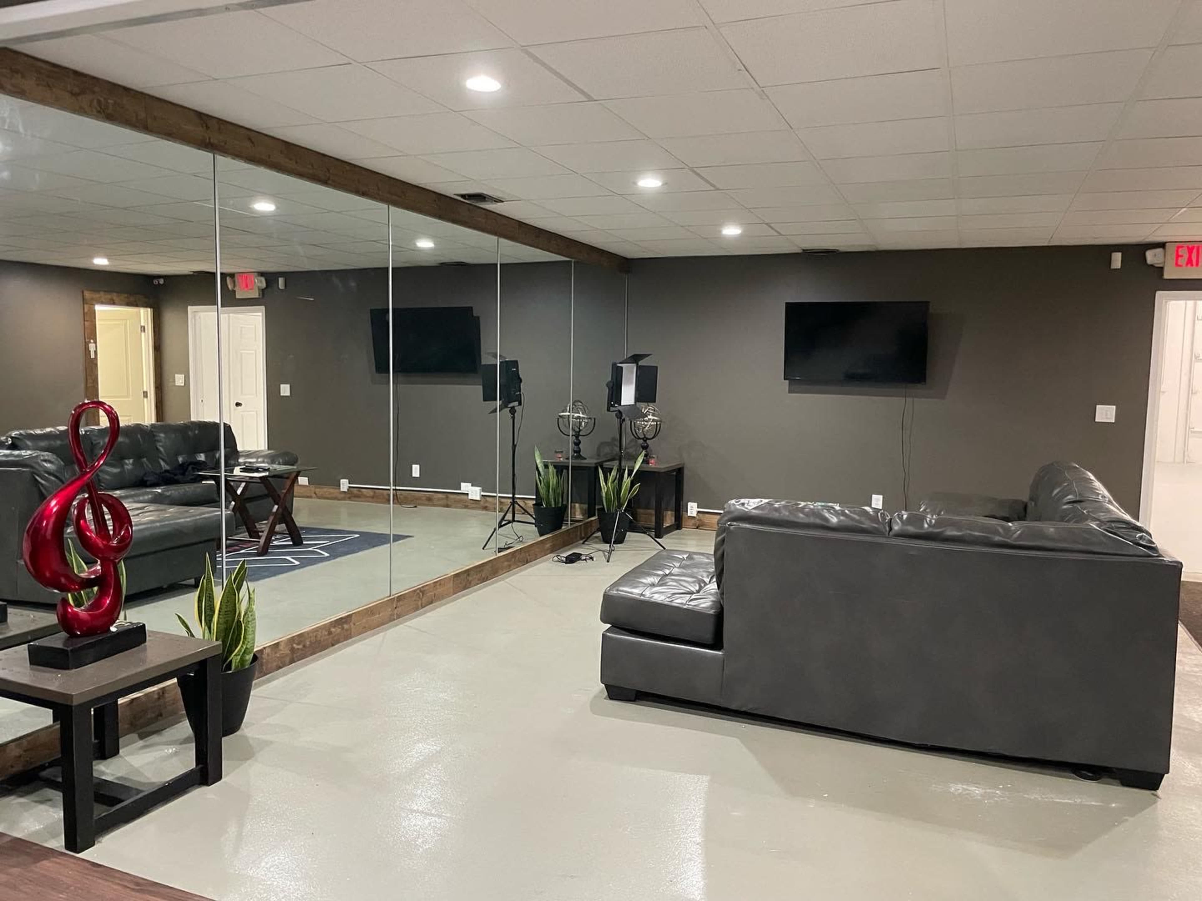 The room features a mirrored wall reflecting two black sofas, a small table with a decorative piece, and two television screens mounted on the walls.