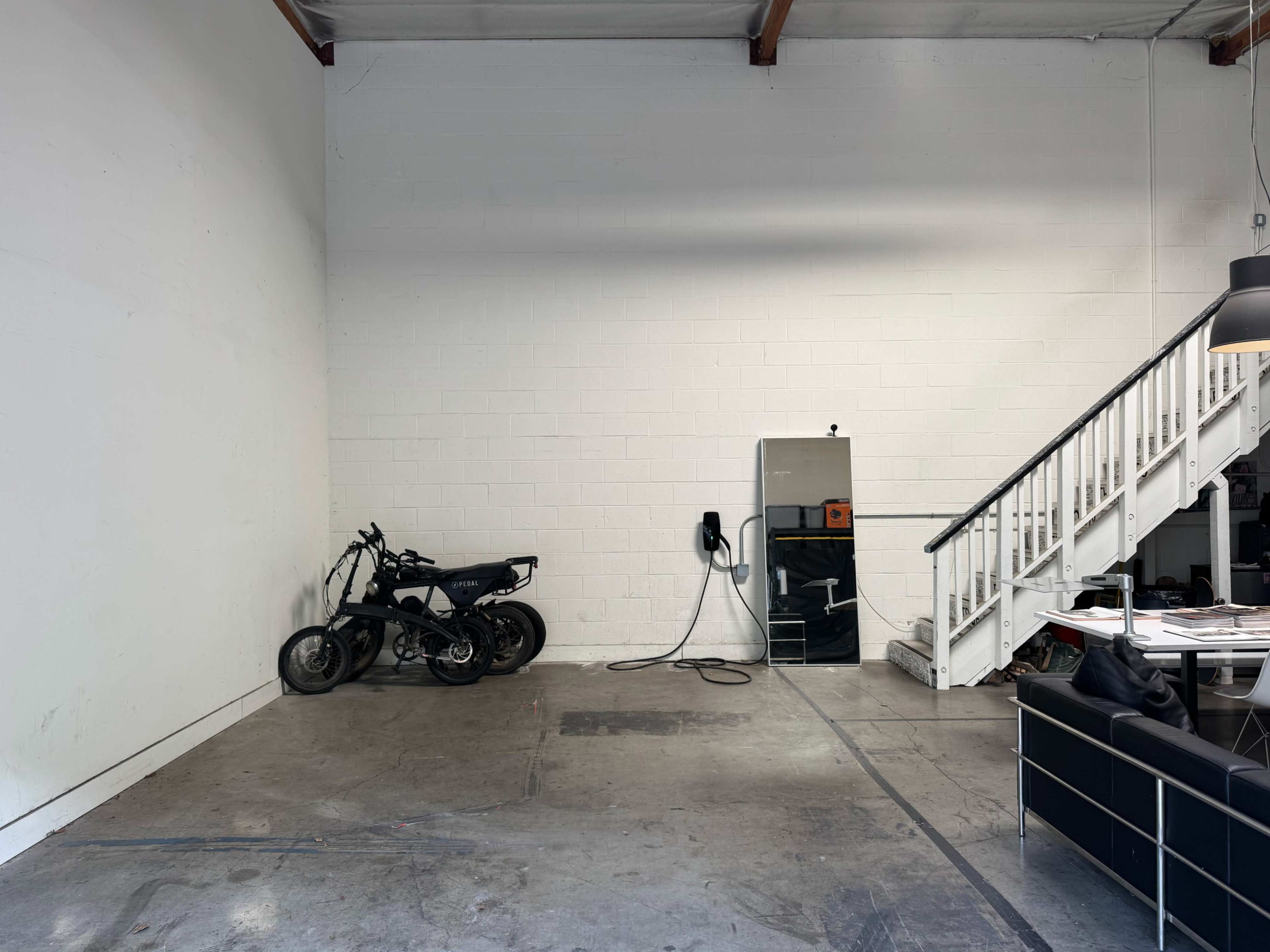 The image shows an empty warehouse space with a motorcycle parked against a white wall, a mirror hanging nearby, and a staircase in the background.