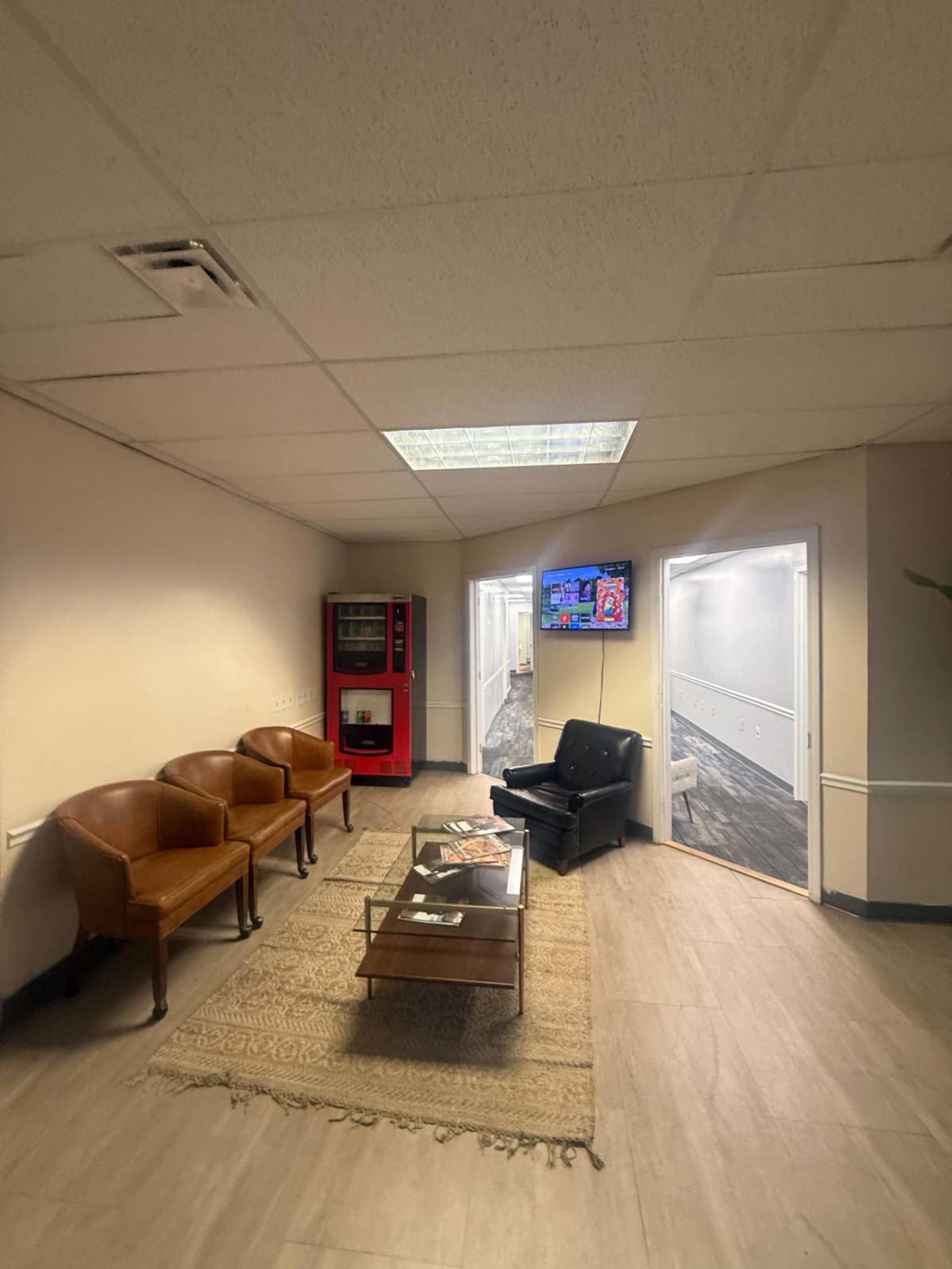 The image shows a waiting area with three brown chairs, a black leather chair, a coffee table on a rug, a vending machine, and a television on the wall, with hallways leading to other rooms in the background.