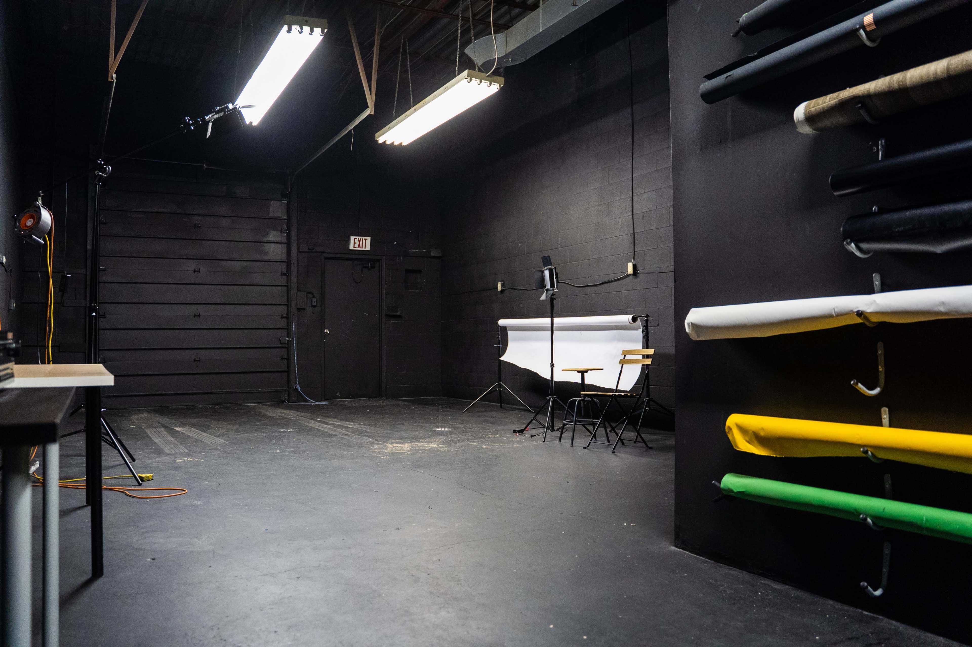 A spacious studio with a black backdrop, a white backdrop support, two chairs, and rolls of colored paper on the wall.