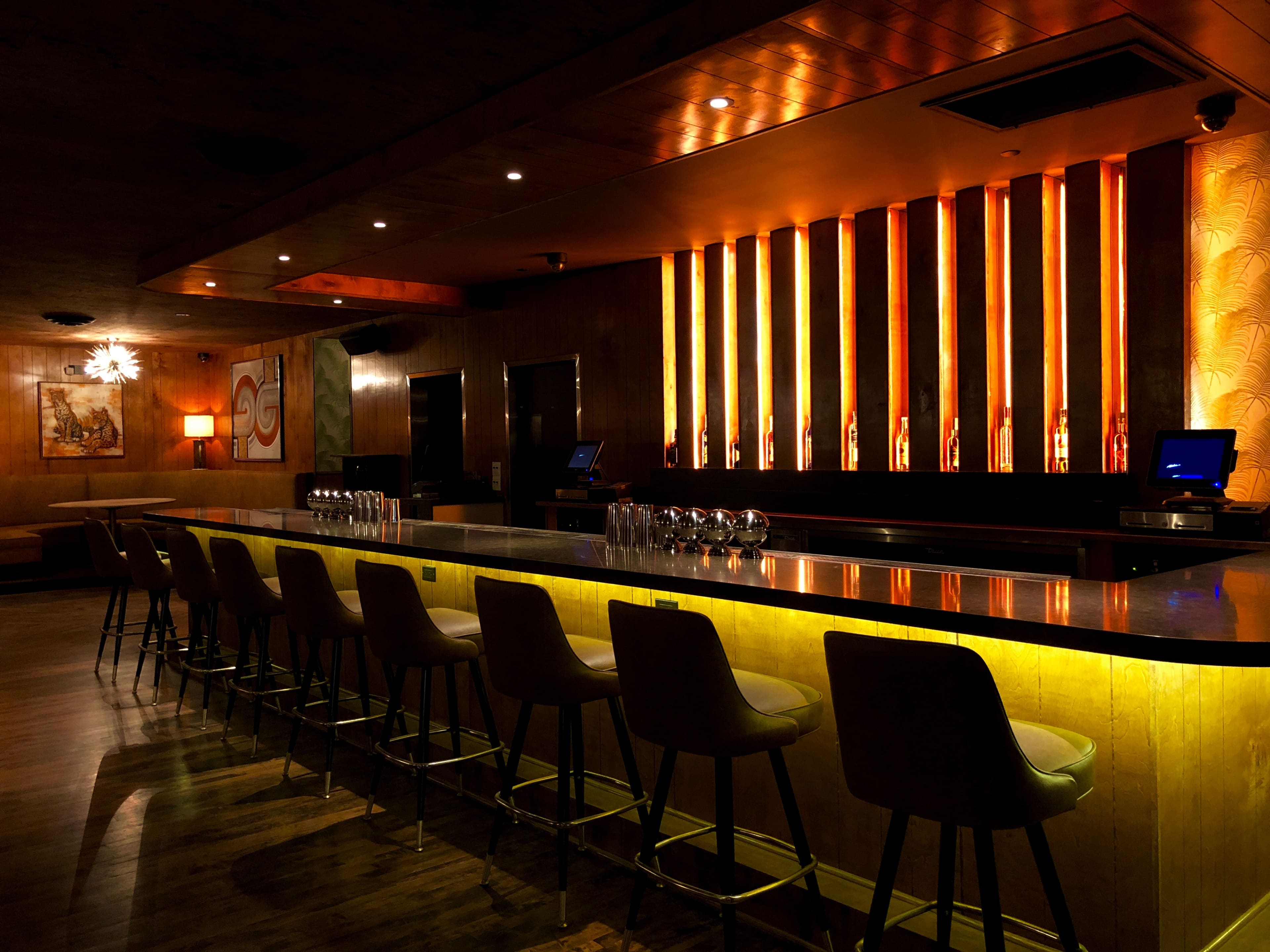 A sleek bar with high stools and illuminated orange vertical panels runs along one side of a dimly lit room.
