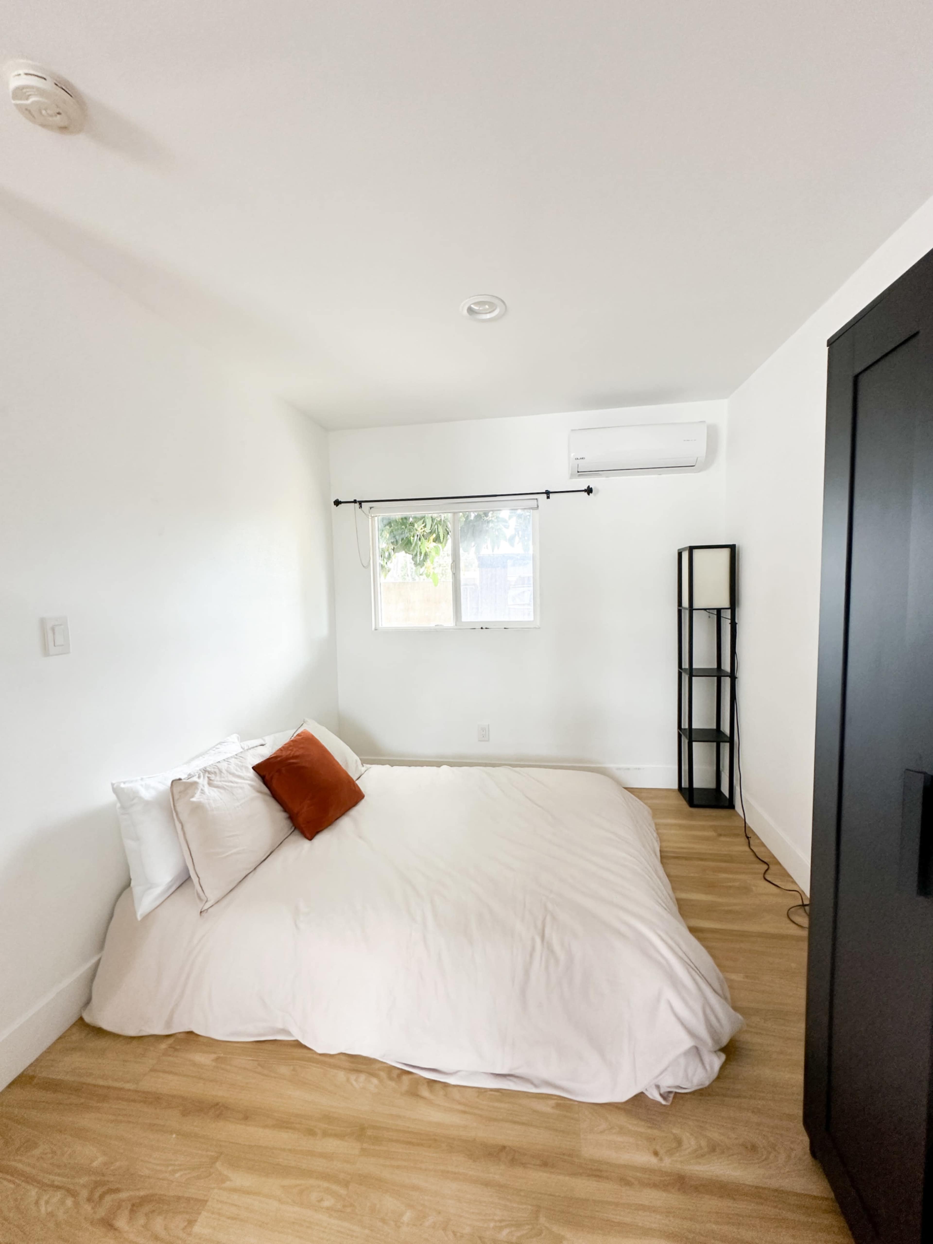 A minimalist bedroom with a low bed on a wooden floor, a window with curtains, and a tall, dark wardrobe.