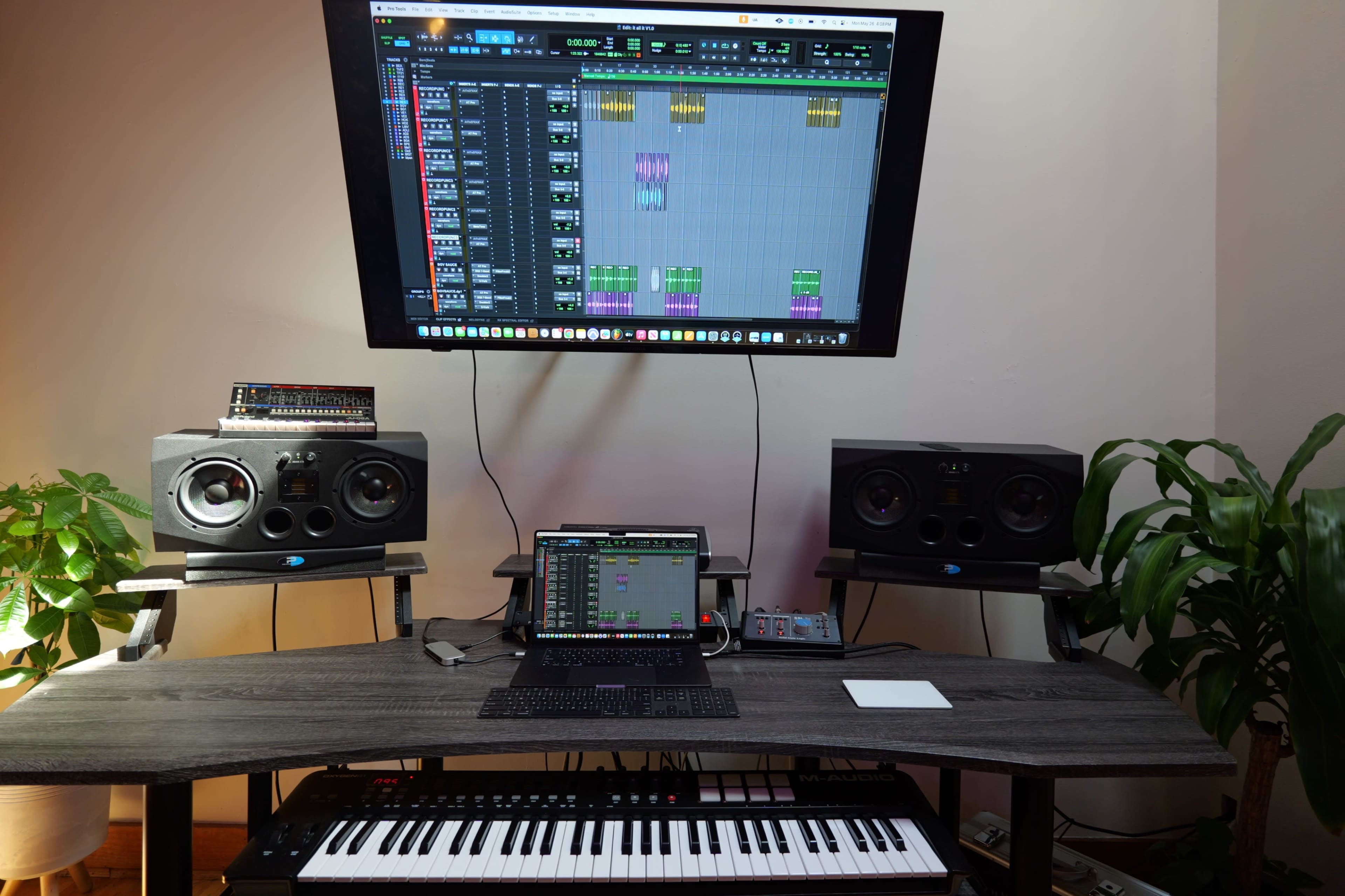 A home music production setup features a computer monitor displaying audio editing software, a keyboard, studio speakers on stands, and a tablet on the desk.