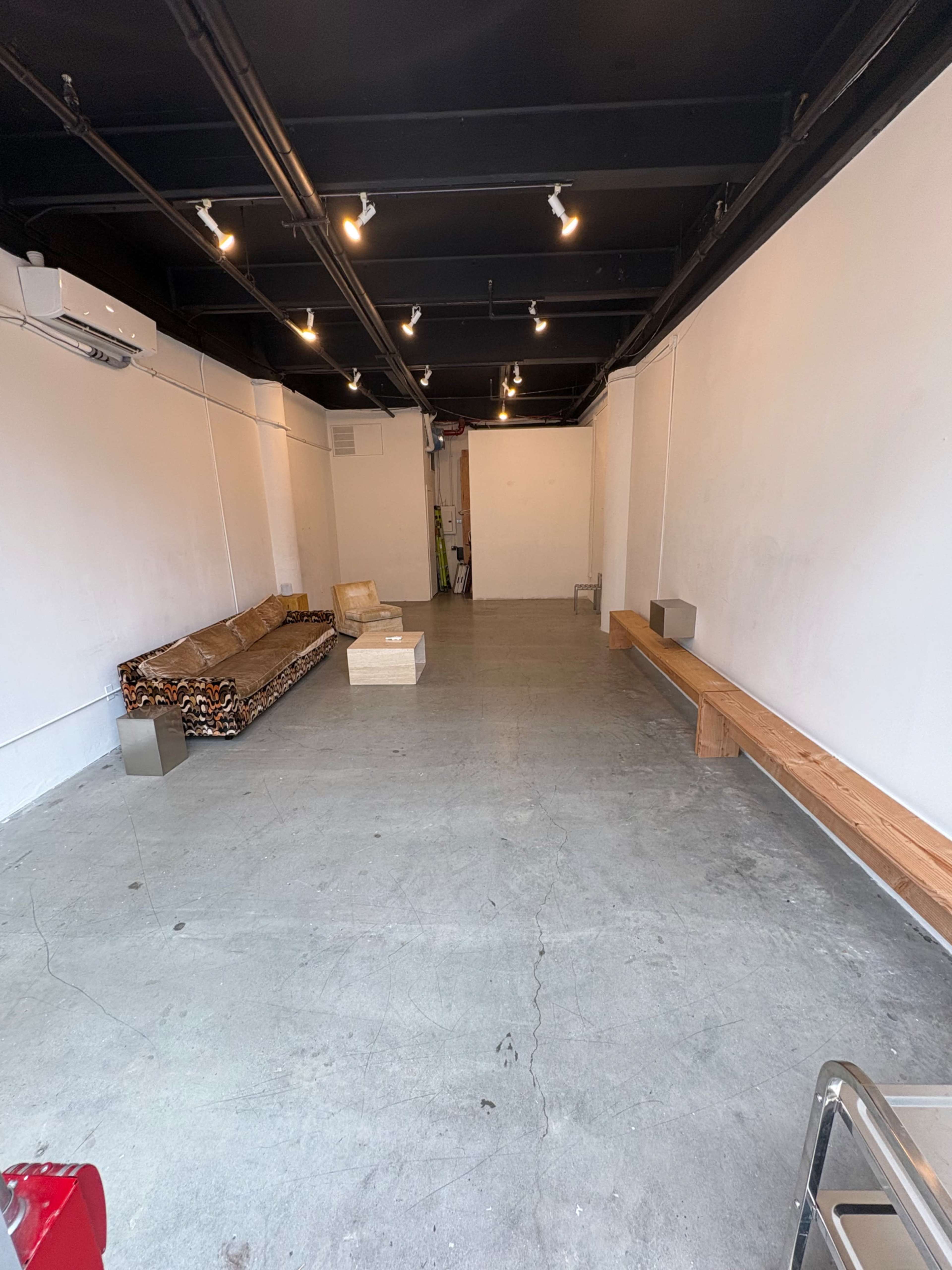 The image shows a spacious, empty room with concrete floors, a patterned couch on one side, and wooden benches along the walls.