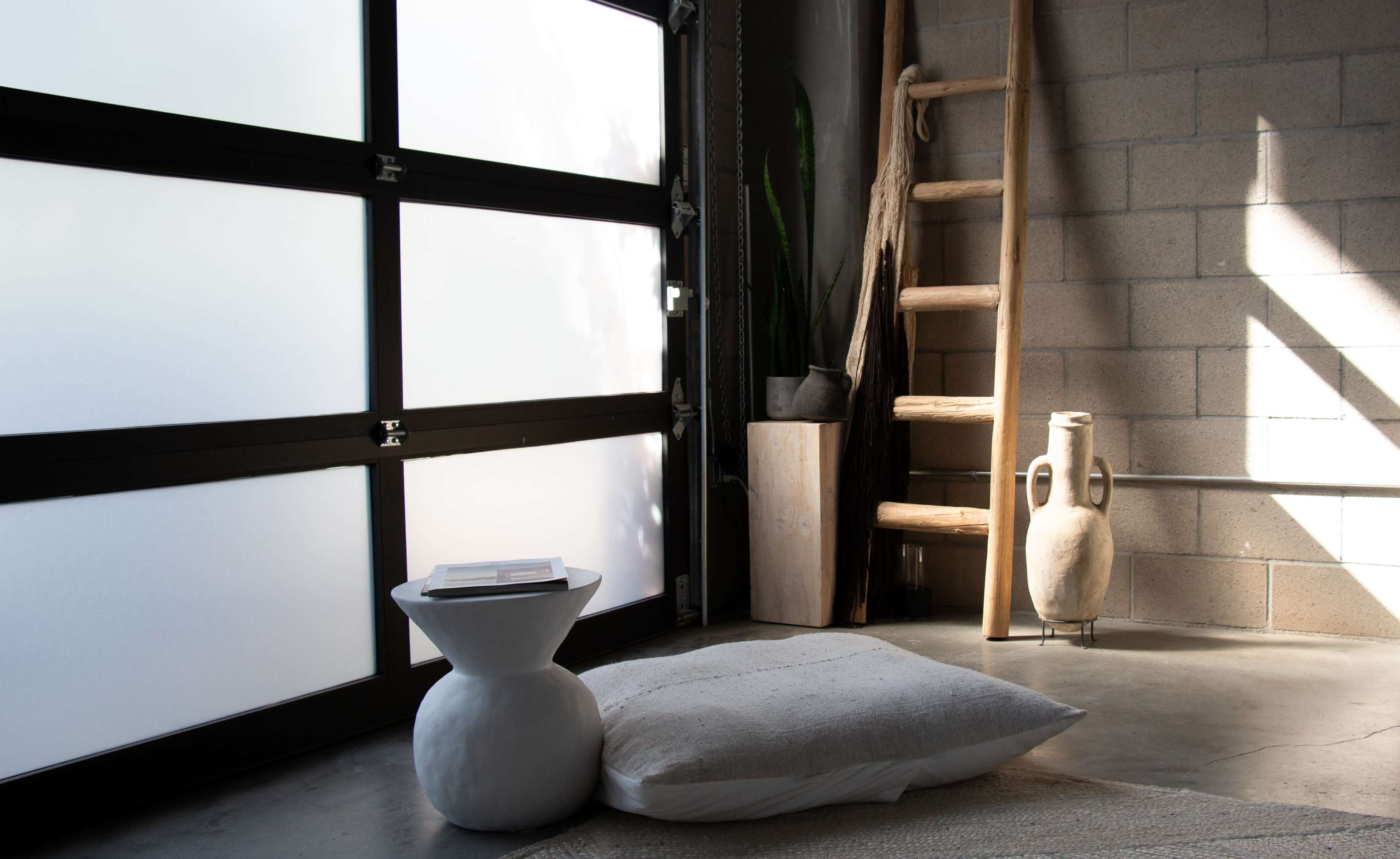A minimalist interior with a concrete floor, a large glass garage door, a wooden ladder, a decorative vase, and a large floor pillow.