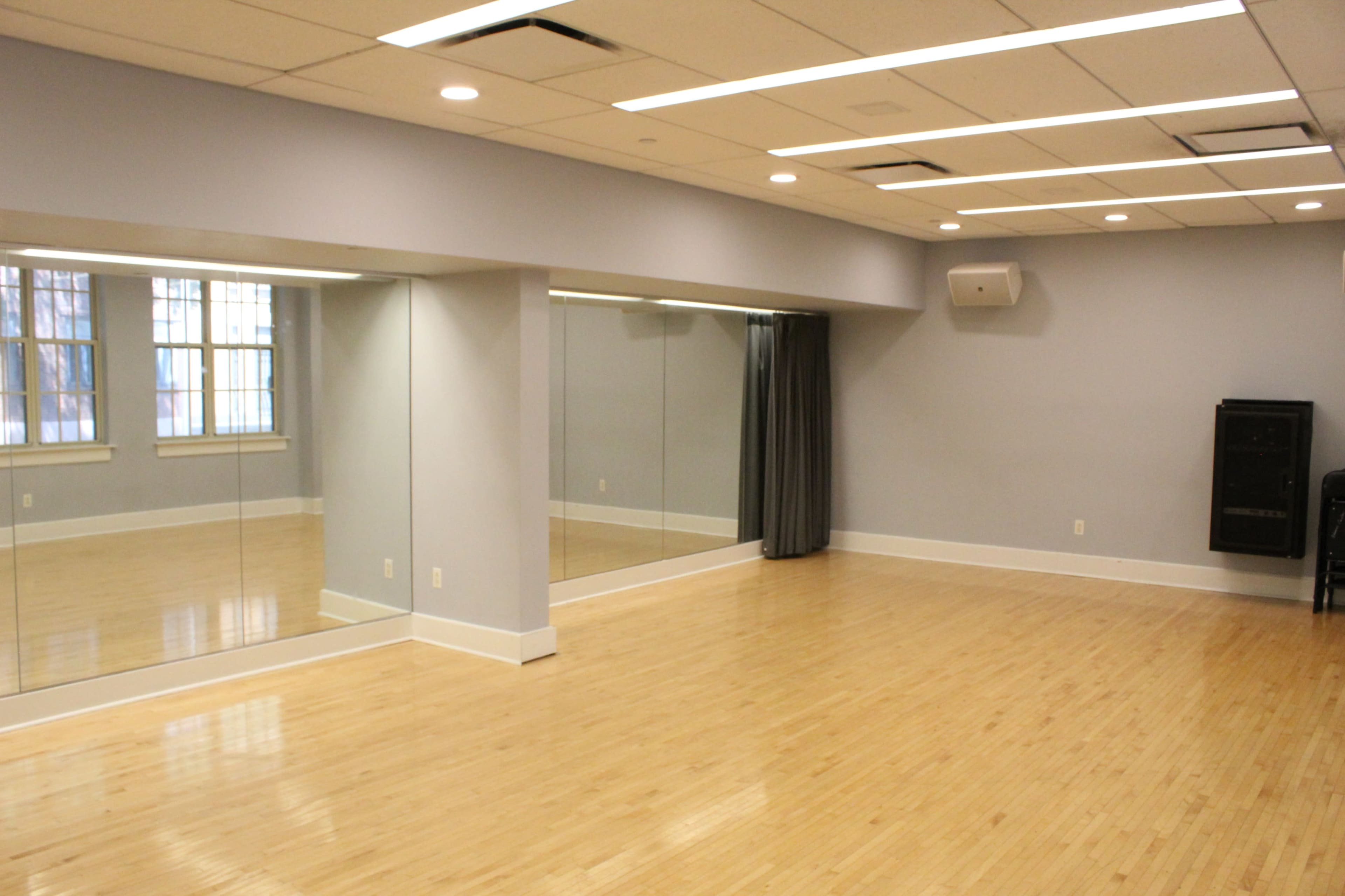 The image shows a bright and spacious room with wooden flooring, large mirrors on one wall, and a curtain along another wall.