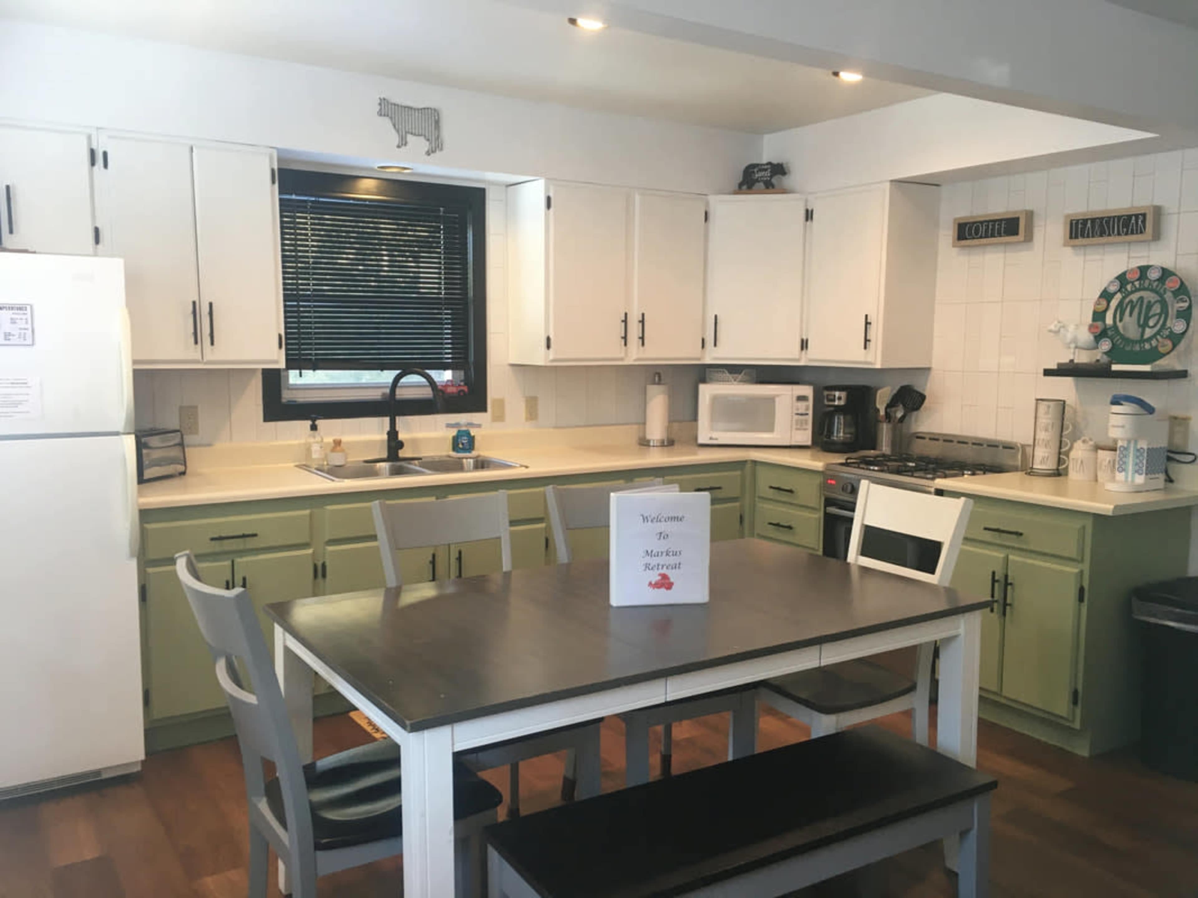 A bright kitchen features white and green cabinetry, a table with seating for six, and modern appliances including a microwave and stove.