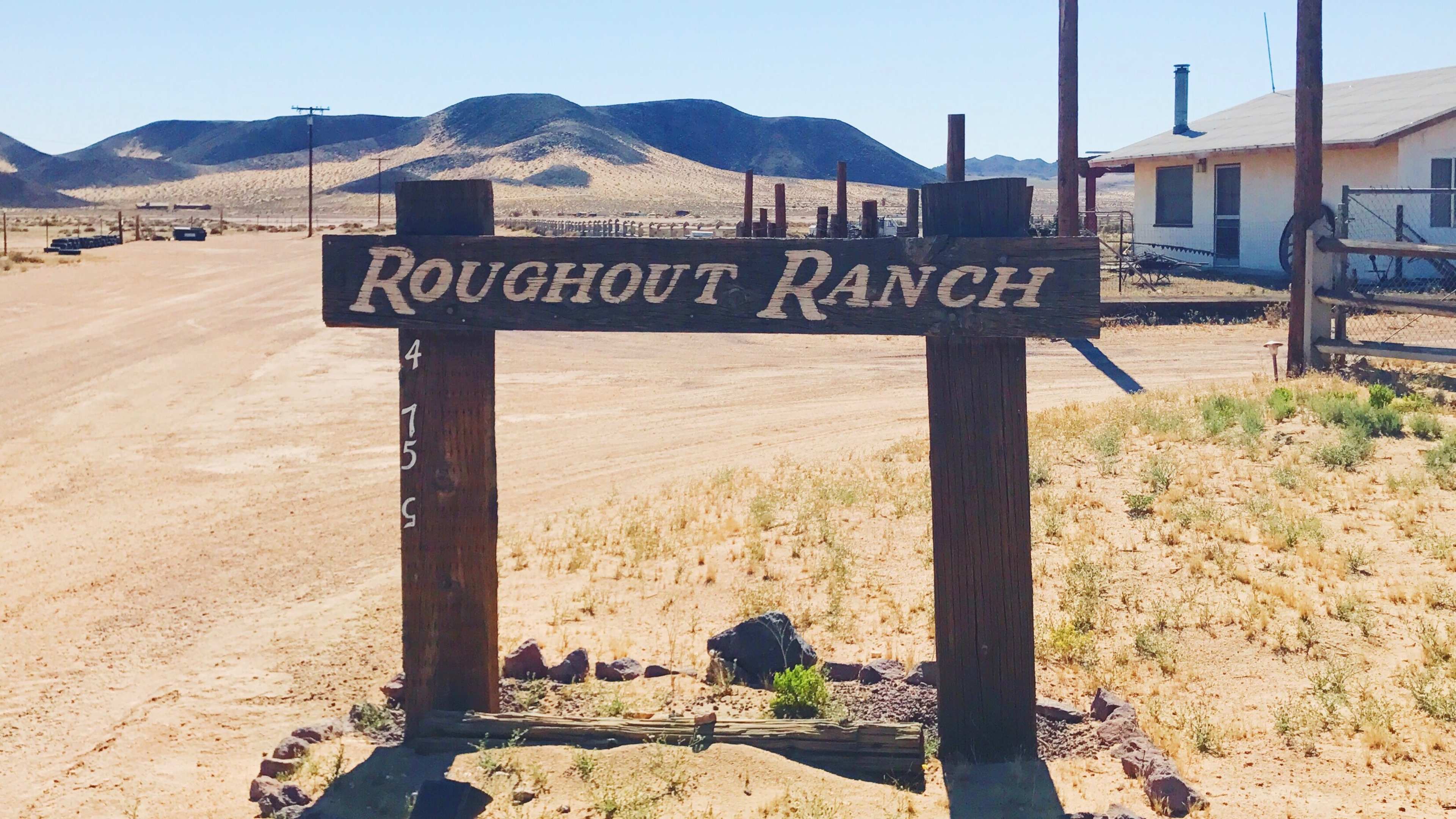 A wooden sign reading "Roughout Ranch" stands at the entrance to a dirt road surrounded by desert landscape and distant mountains.
