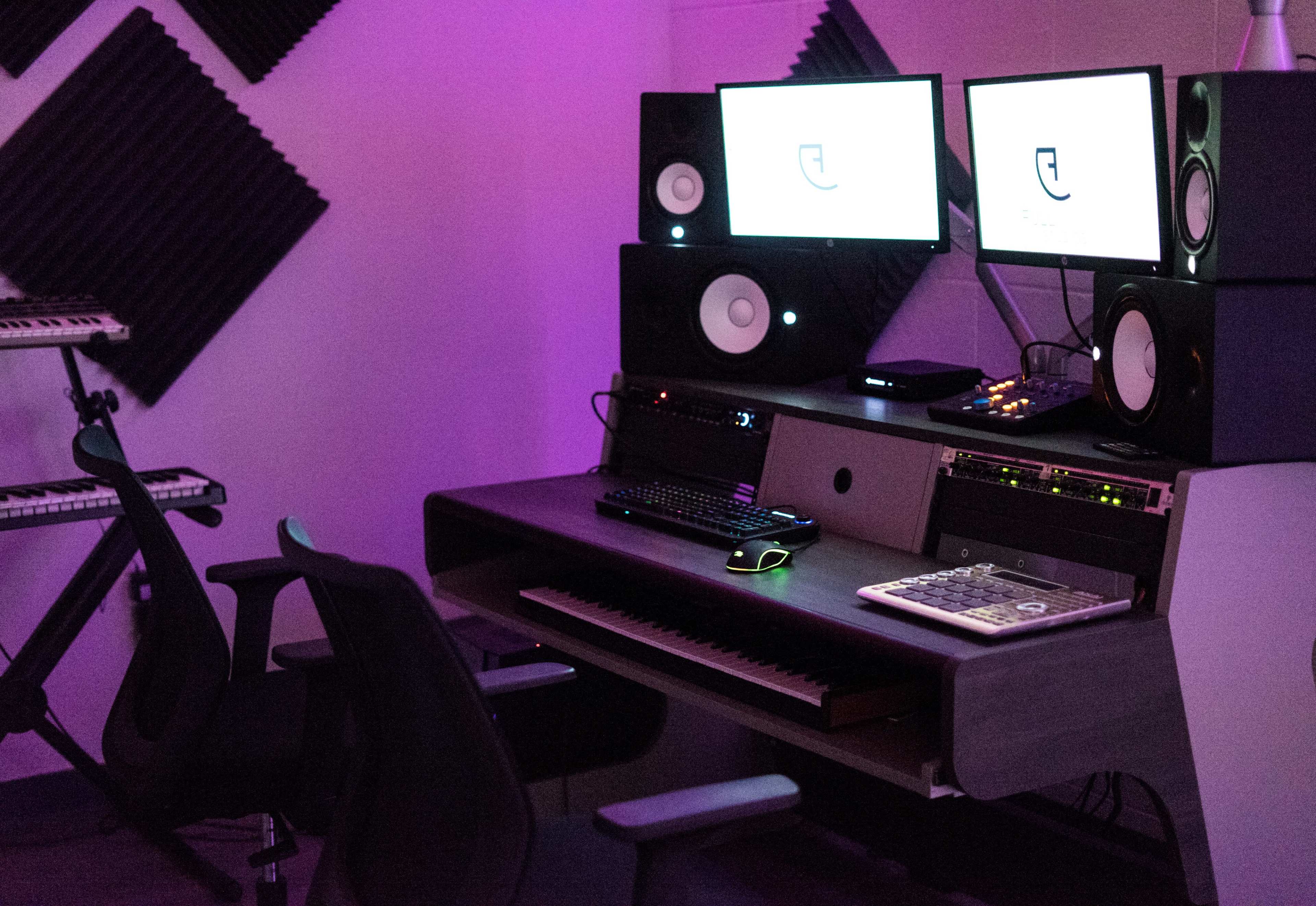 A music production studio with two computer monitors, studio speakers, a keyboard, and a mixing console, set against soundproofing panels.